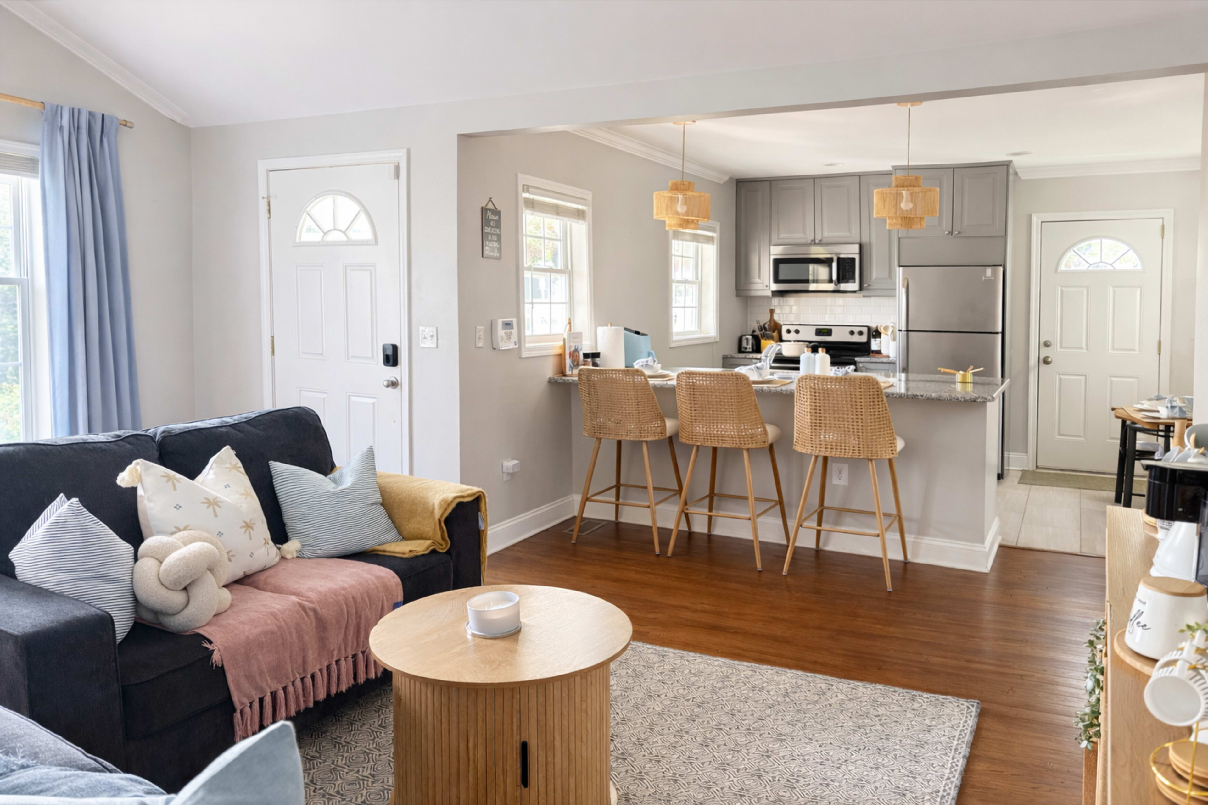 The image shows a cozy living area with a navy blue sofa, a round wooden coffee table, and a connected kitchen featuring barstool seating and modern appliances.