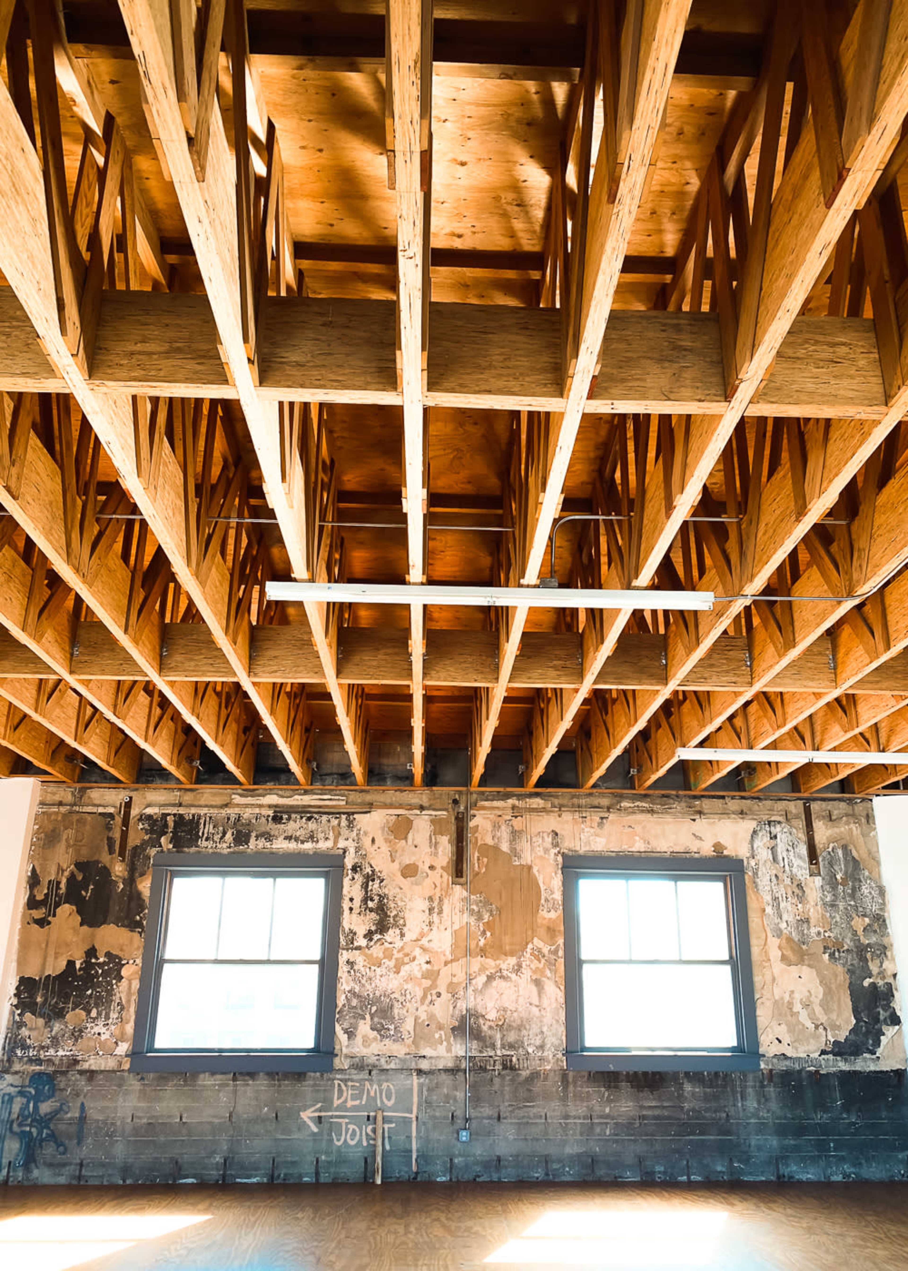 The image shows a spacious room with wooden beams overhead, two windows on one wall, and exposed, weathered wall surfaces.