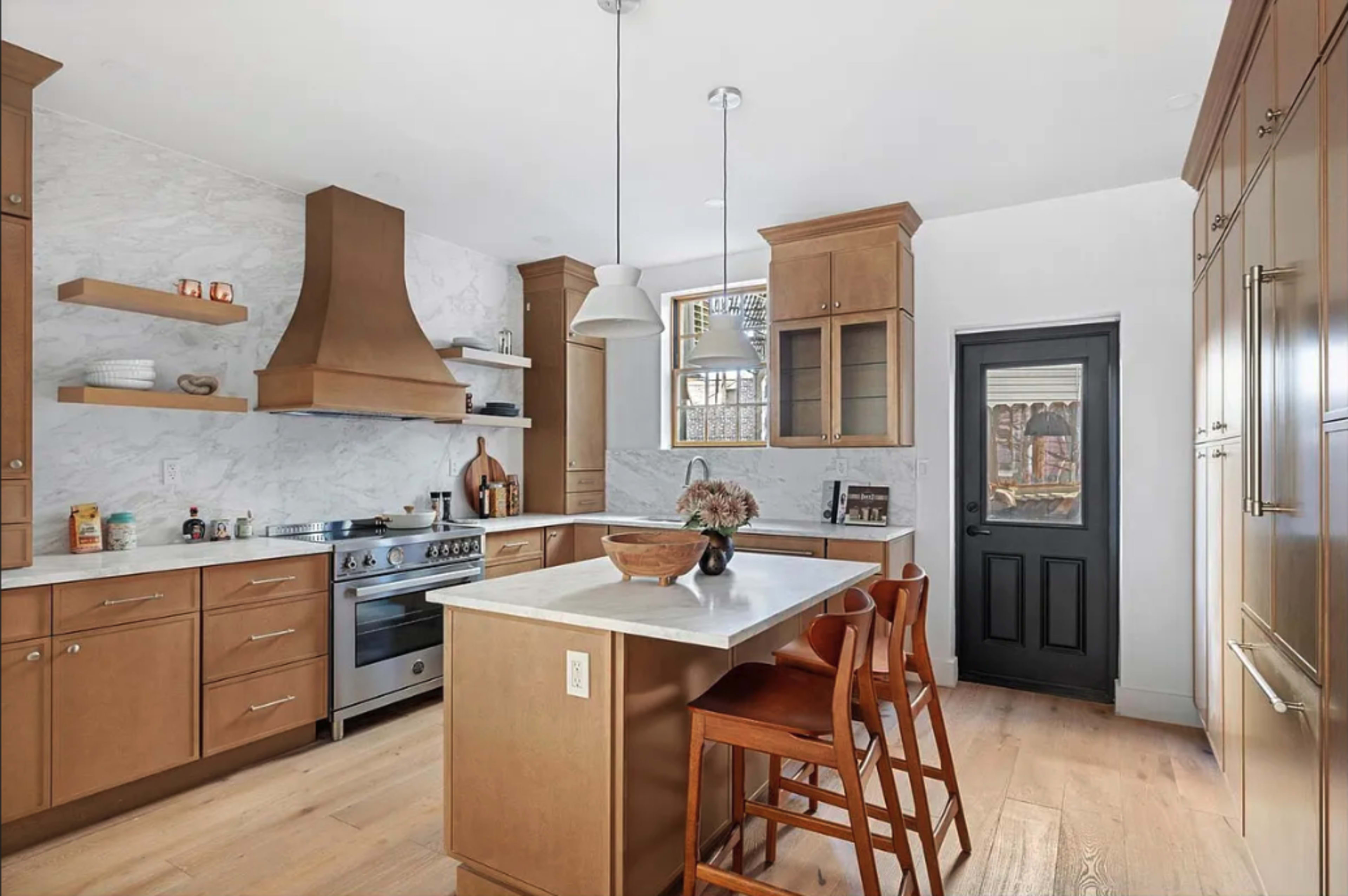 The image shows a modern kitchen featuring wood cabinetry, a stainless steel oven, and a central island with two wooden stools.