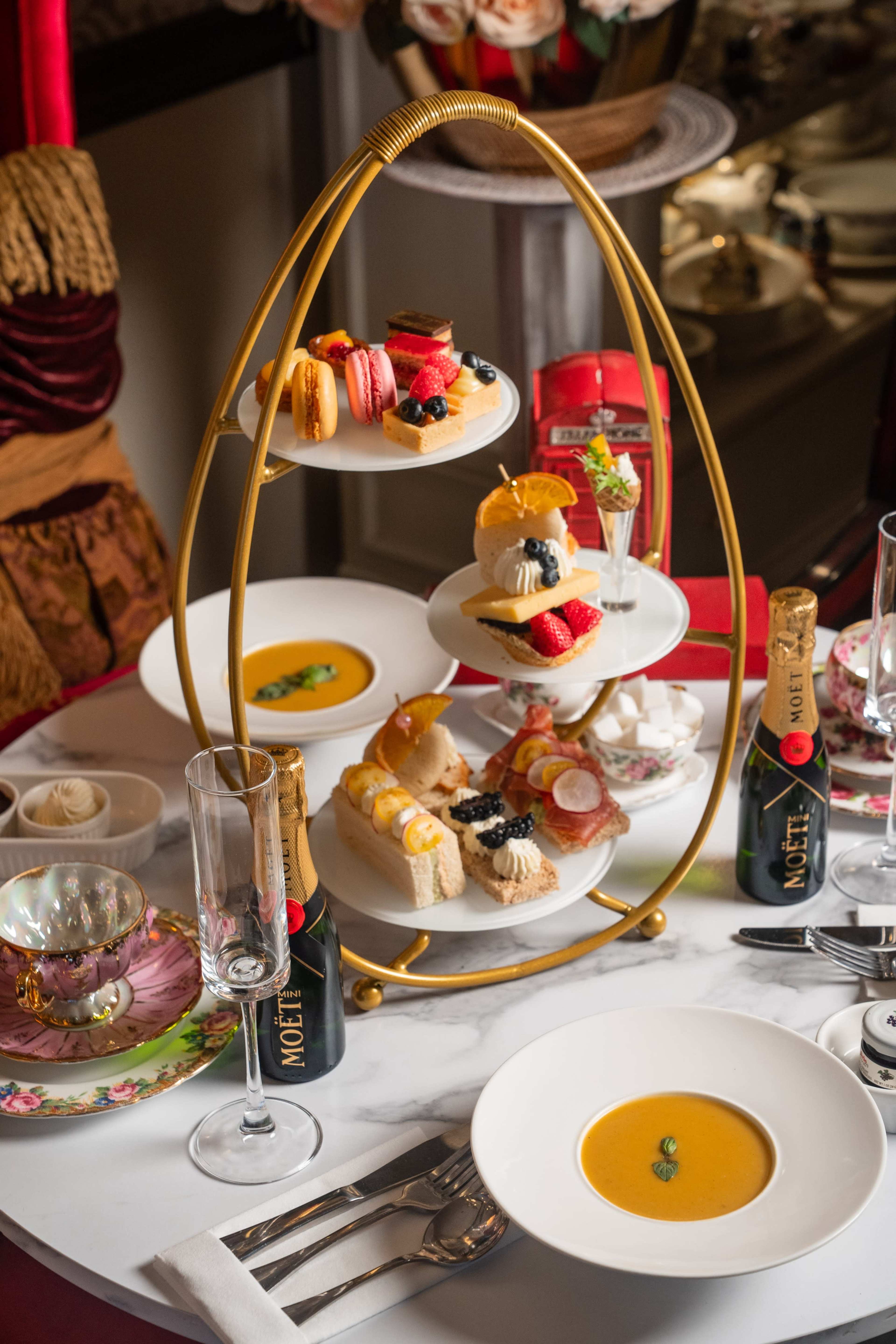 A three-tiered stand displays an assortment of pastries, cakes, and finger foods alongside champagne flutes and bowls of soup on a marble table.