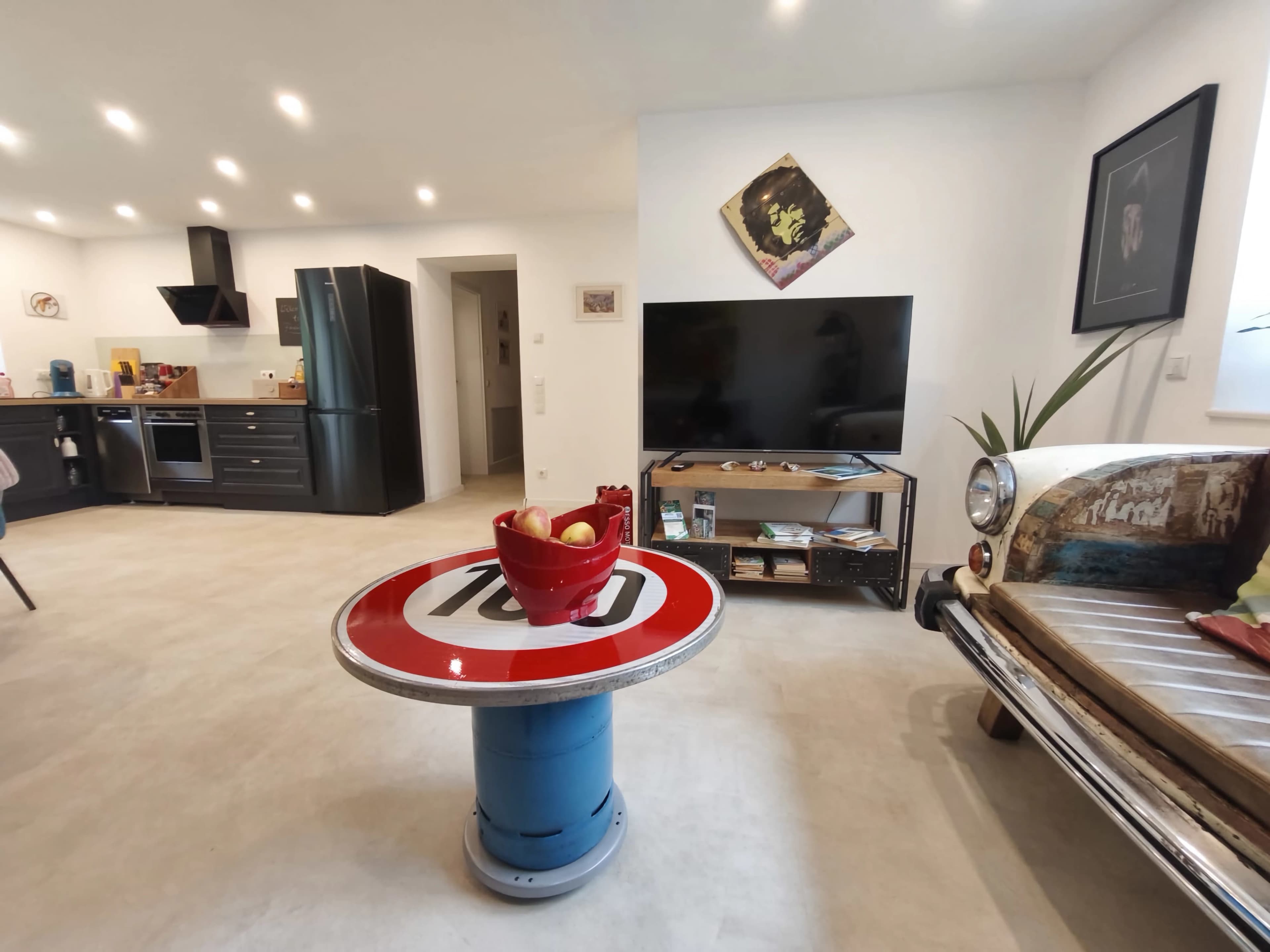 The image shows a modern living space featuring a kitchen area, a black refrigerator, a television, a coffee table made from a circular traffic sign, and a vintage car seat as seating.