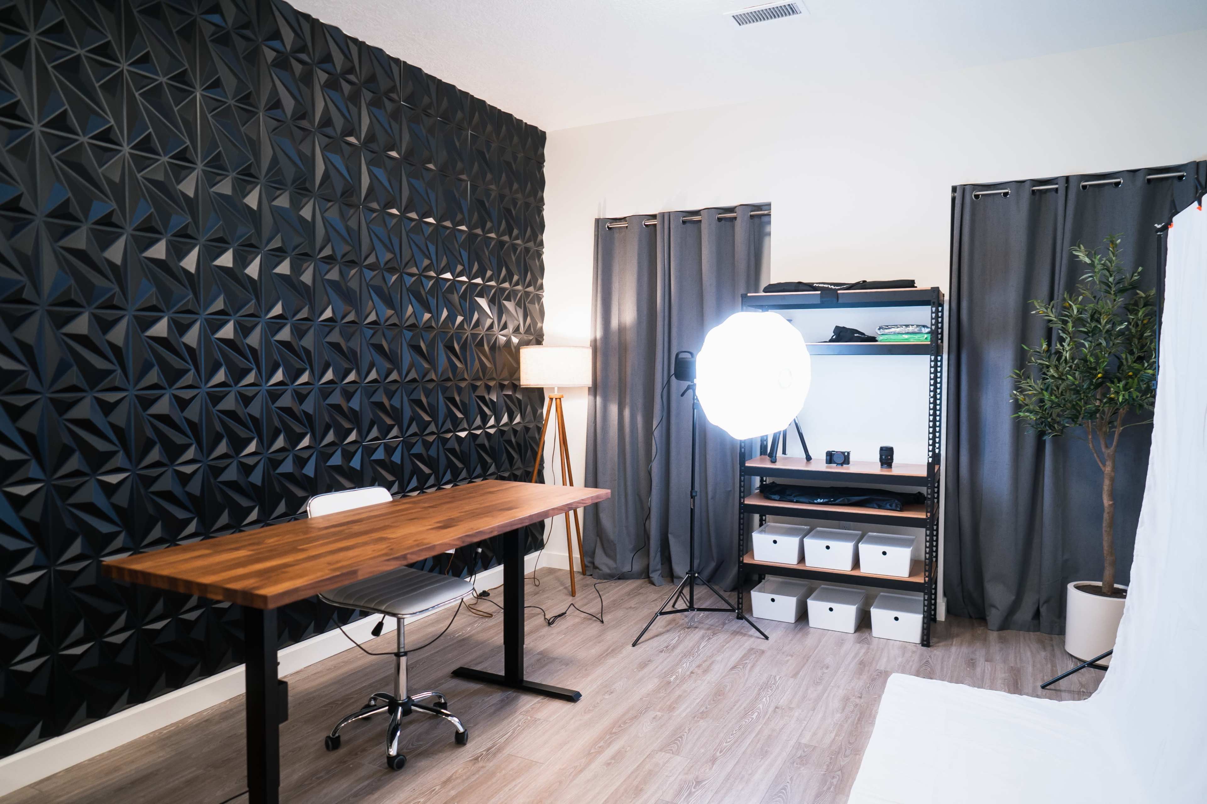 A minimalist office space featuring a wooden desk, a large light source, storage boxes, and a textured black wall.