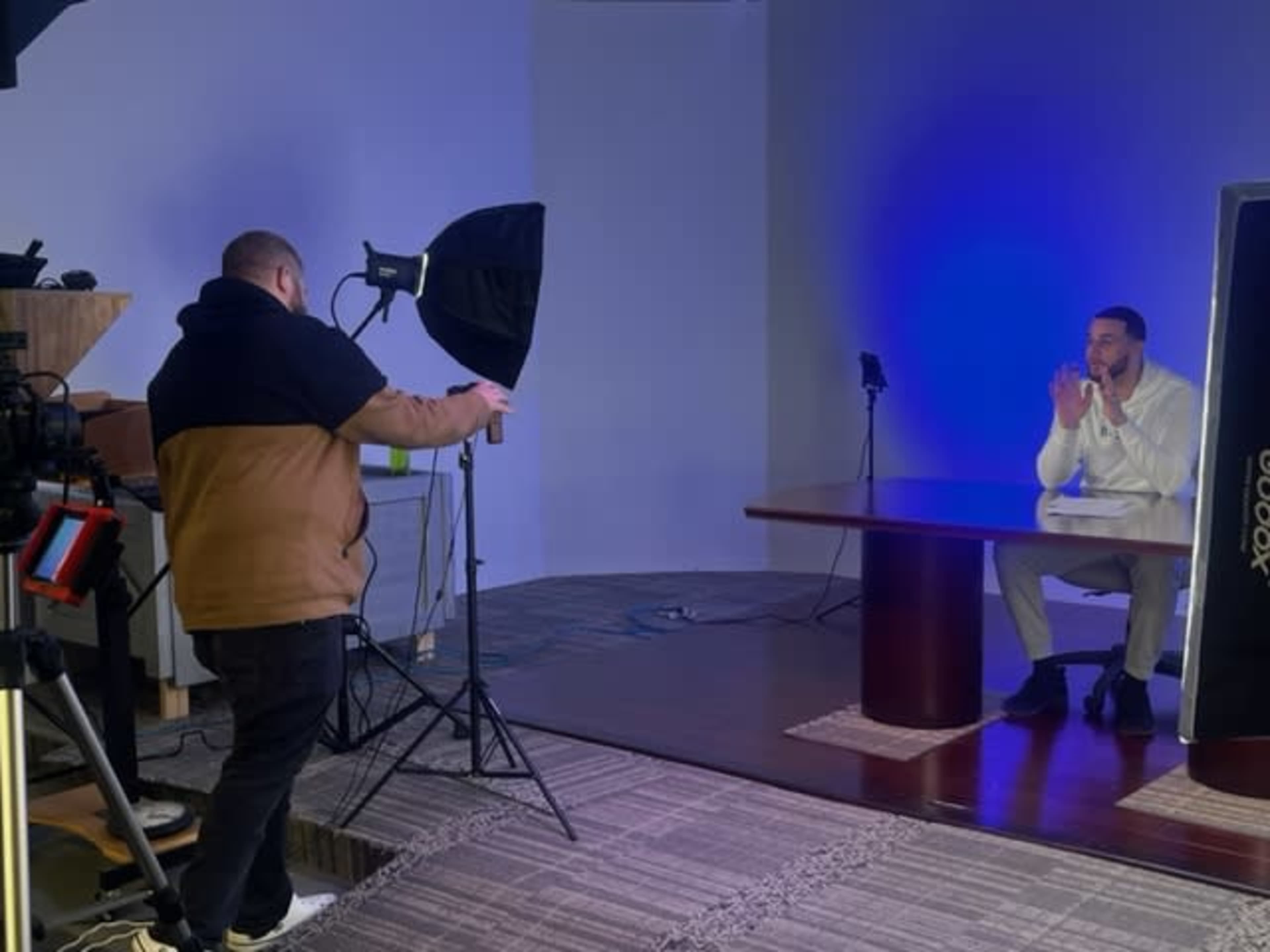 A man sits at a table in a studio while another man adjusts a light setup and camera equipment.