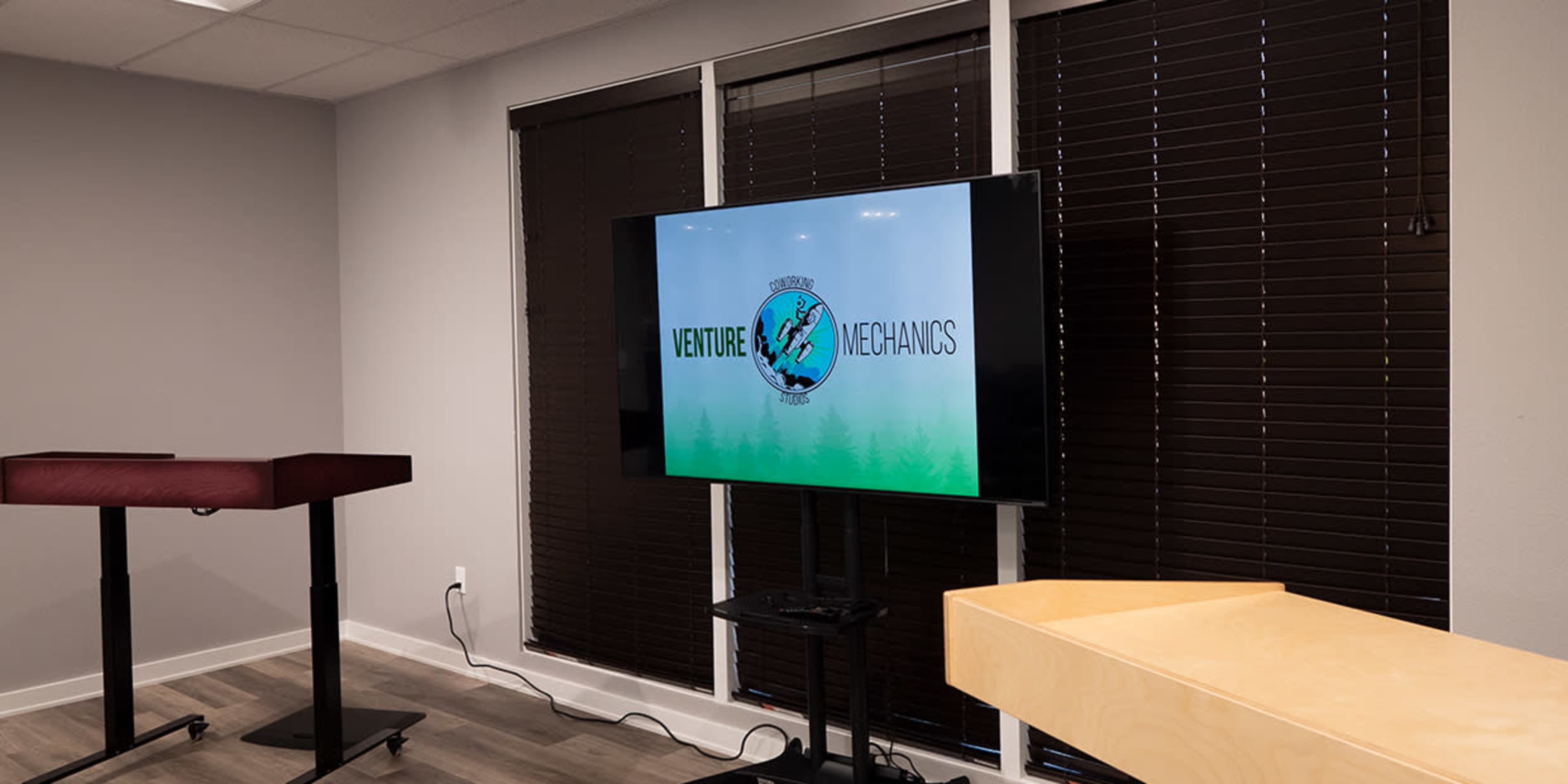 A television display shows a logo for "Venture Mechanos" in a modern room with dark blinds and minimalist furniture.