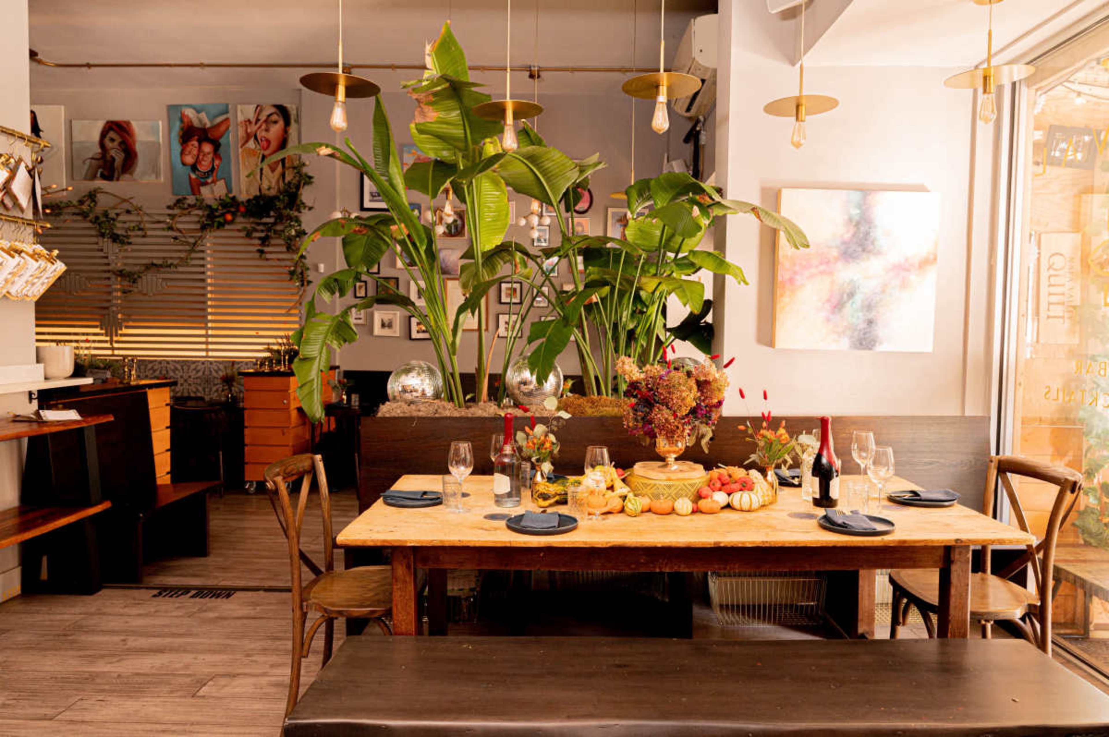 The image shows a stylish restaurant interior with a long wooden table set for dining, surrounded by greenery and decorated with artwork on the walls.