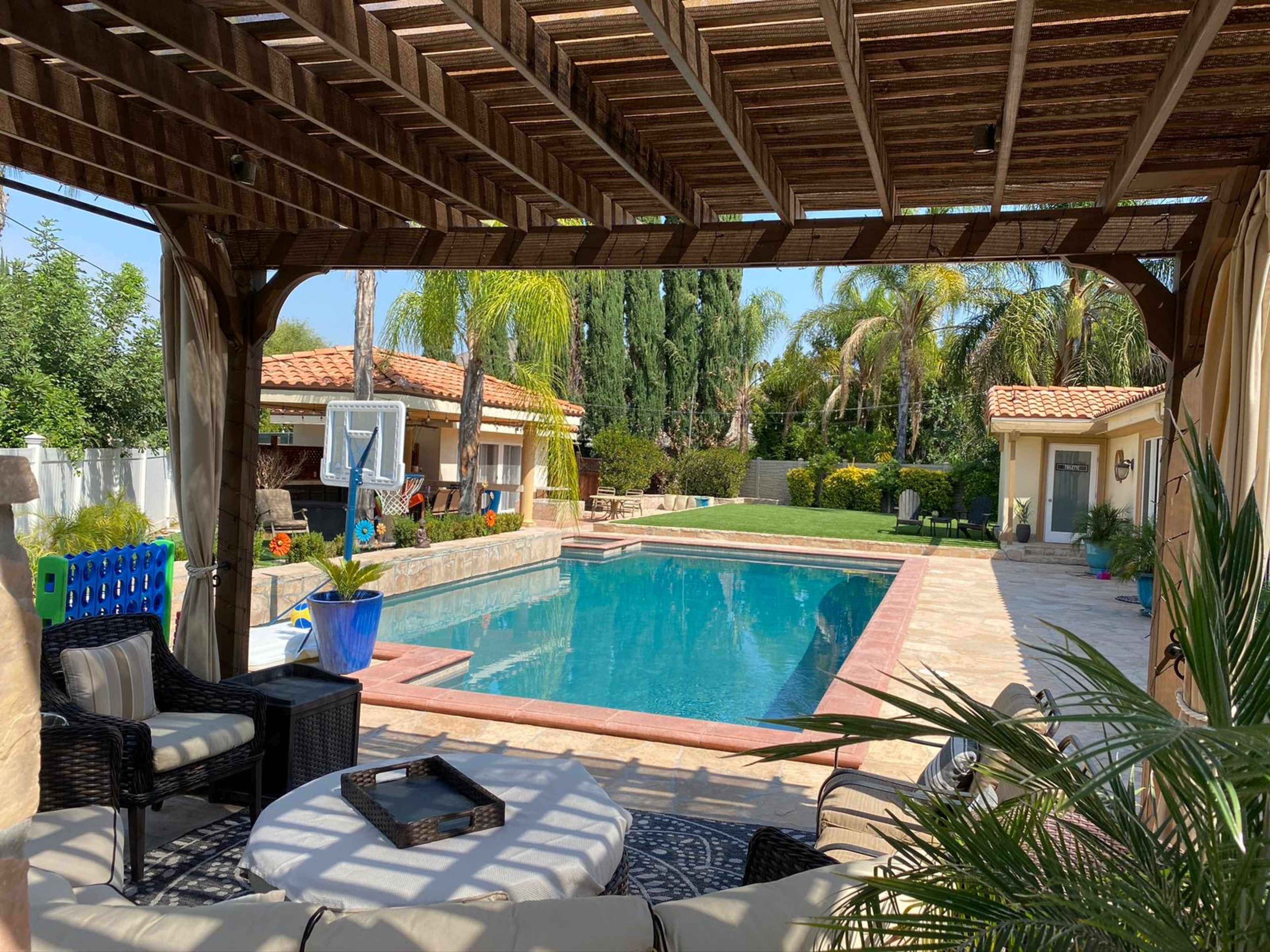 A pool area surrounded by tropical plants and a covered patio with seating, leading to a well-maintained lawn and villa-style buildings.