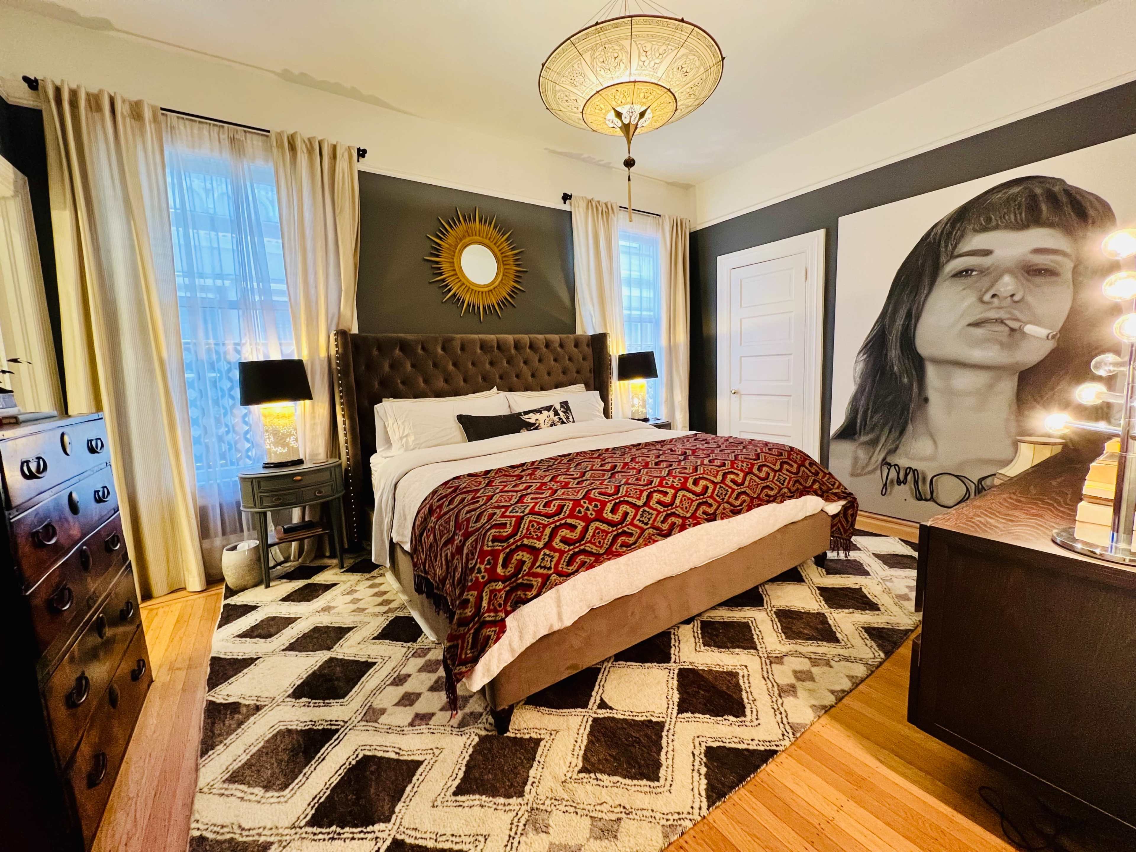 A stylish bedroom featuring a large bed with a patterned throw, two bedside lamps, a wall-mounted mirror, and a prominent mural of a woman.