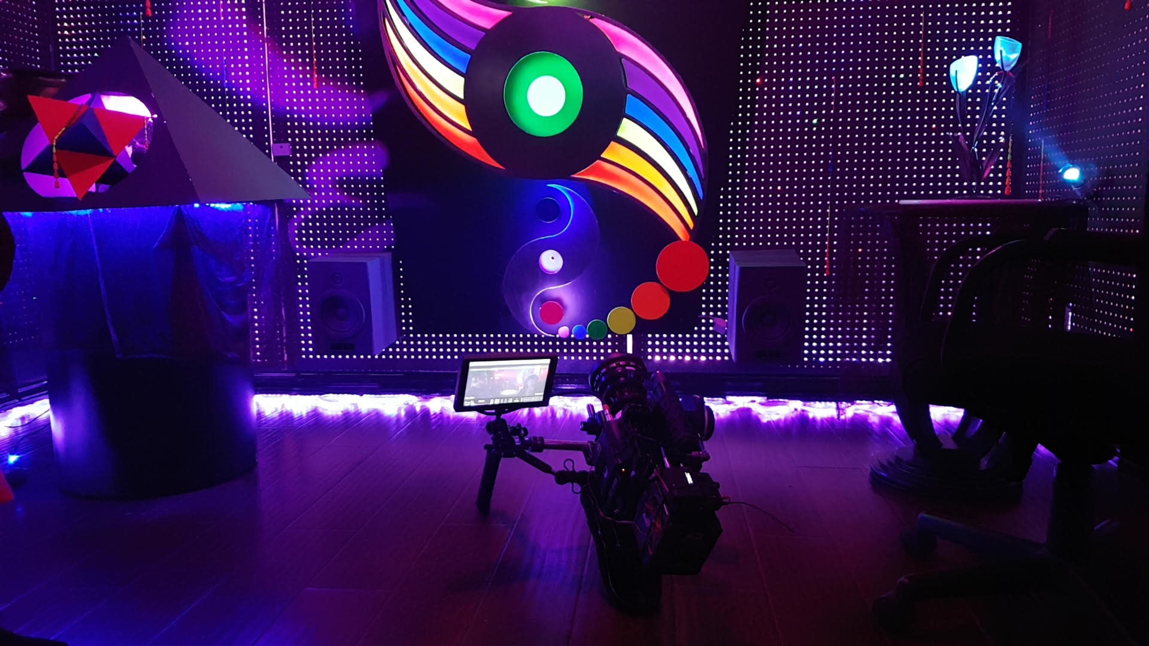 The scene features a colorful illuminated backdrop with circular patterns, a pyramid sculpture, speakers, and a camera set up on a wooden floor.