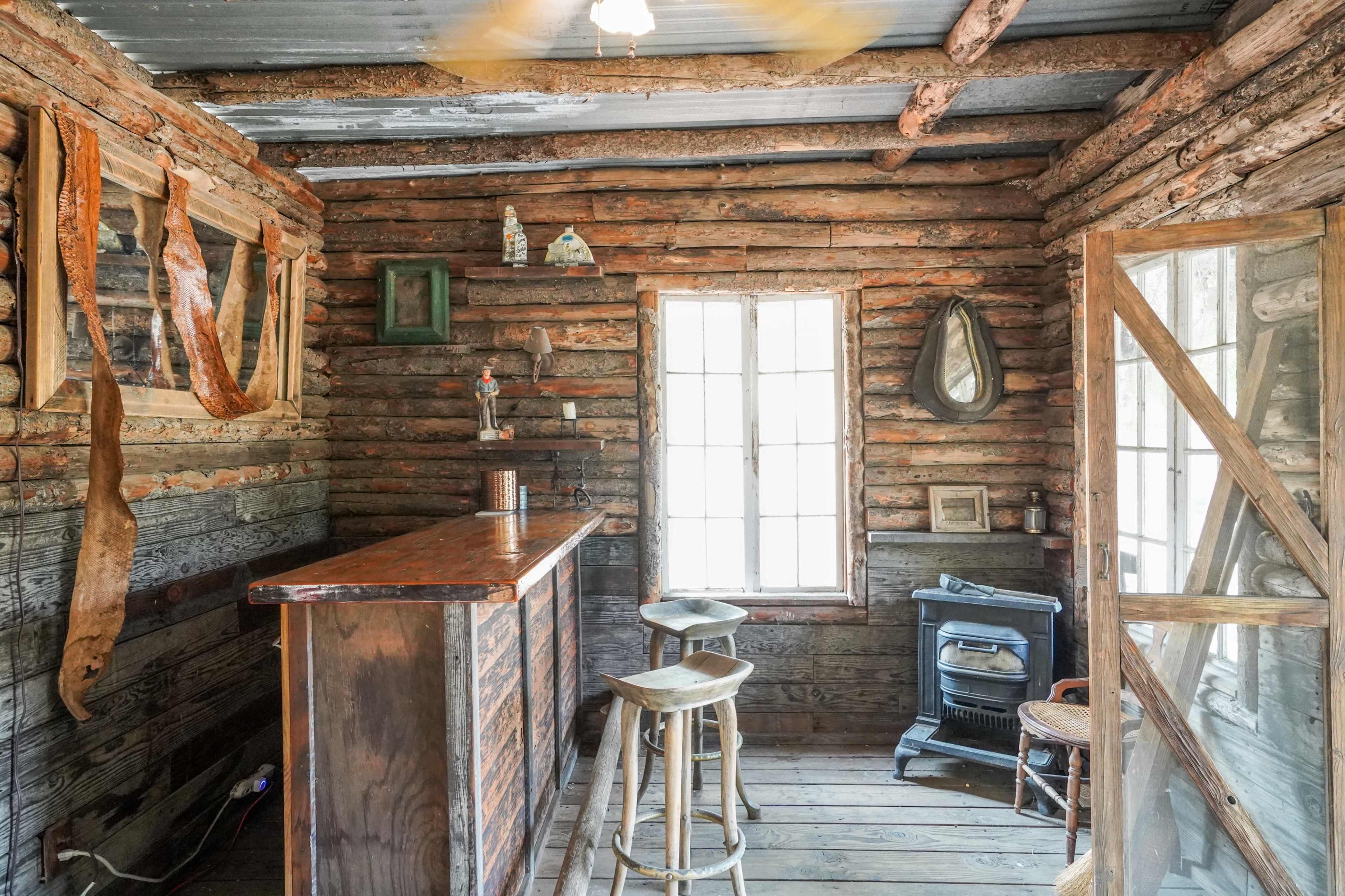 The interior of a rustic cabin features a wooden bar, stools, and a vintage stove surrounded by log walls and windows.