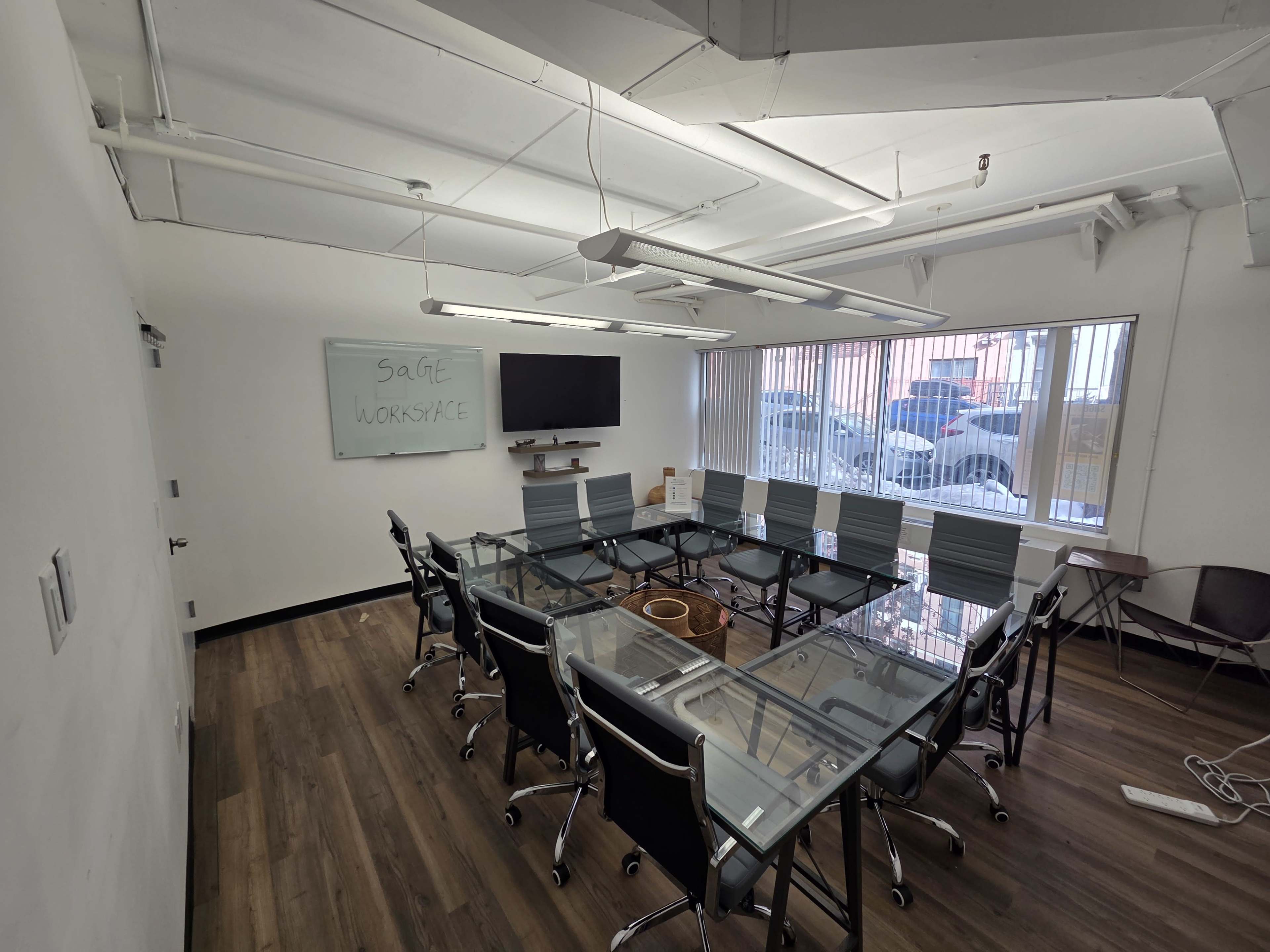 The image shows a modern conference room with a glass-top table surrounded by ergonomic chairs, a wall-mounted TV, and a whiteboard.