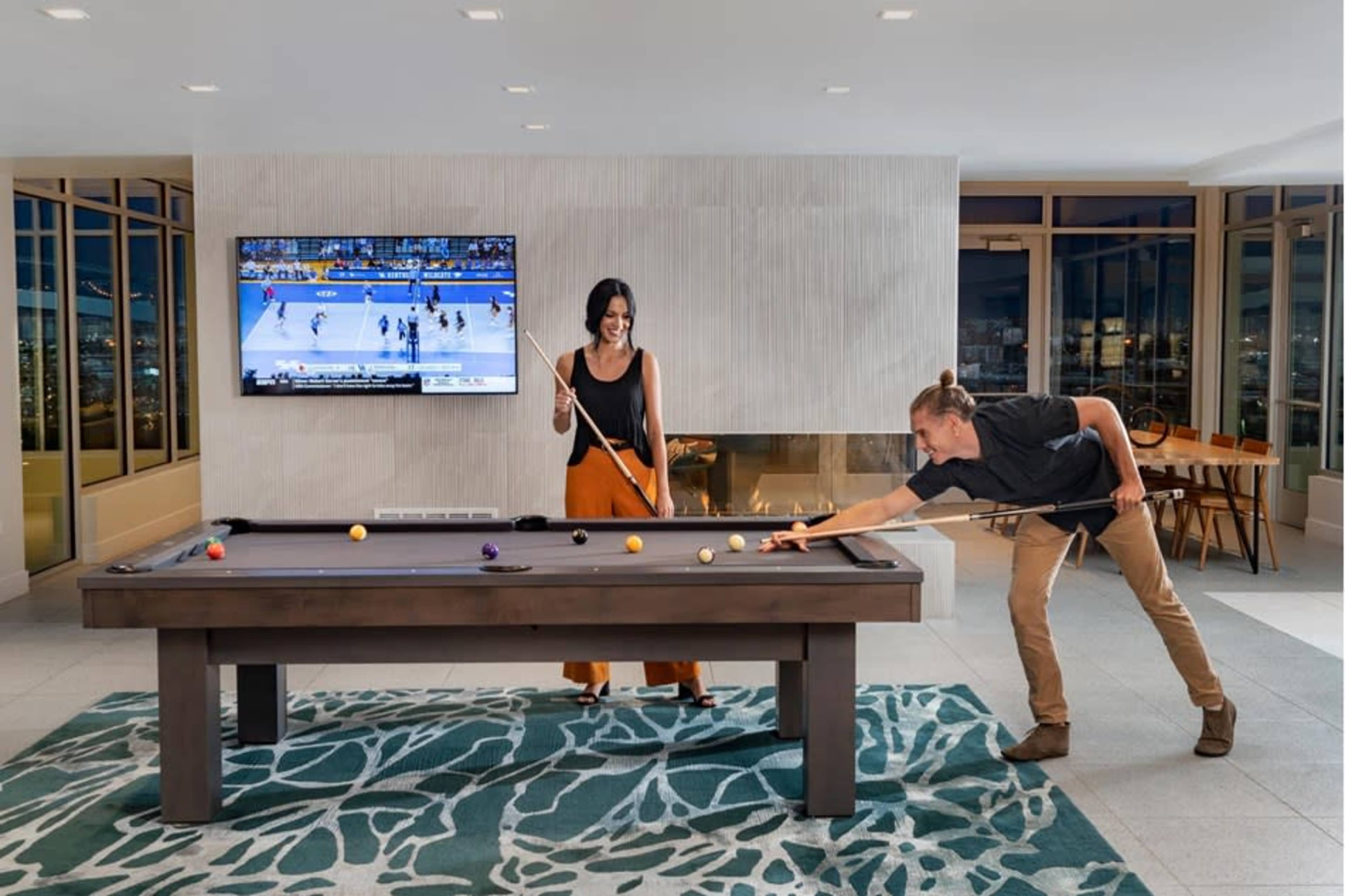 A man and a woman play pool in a brightly lit room with large windows and a television showing a sports game in the background.