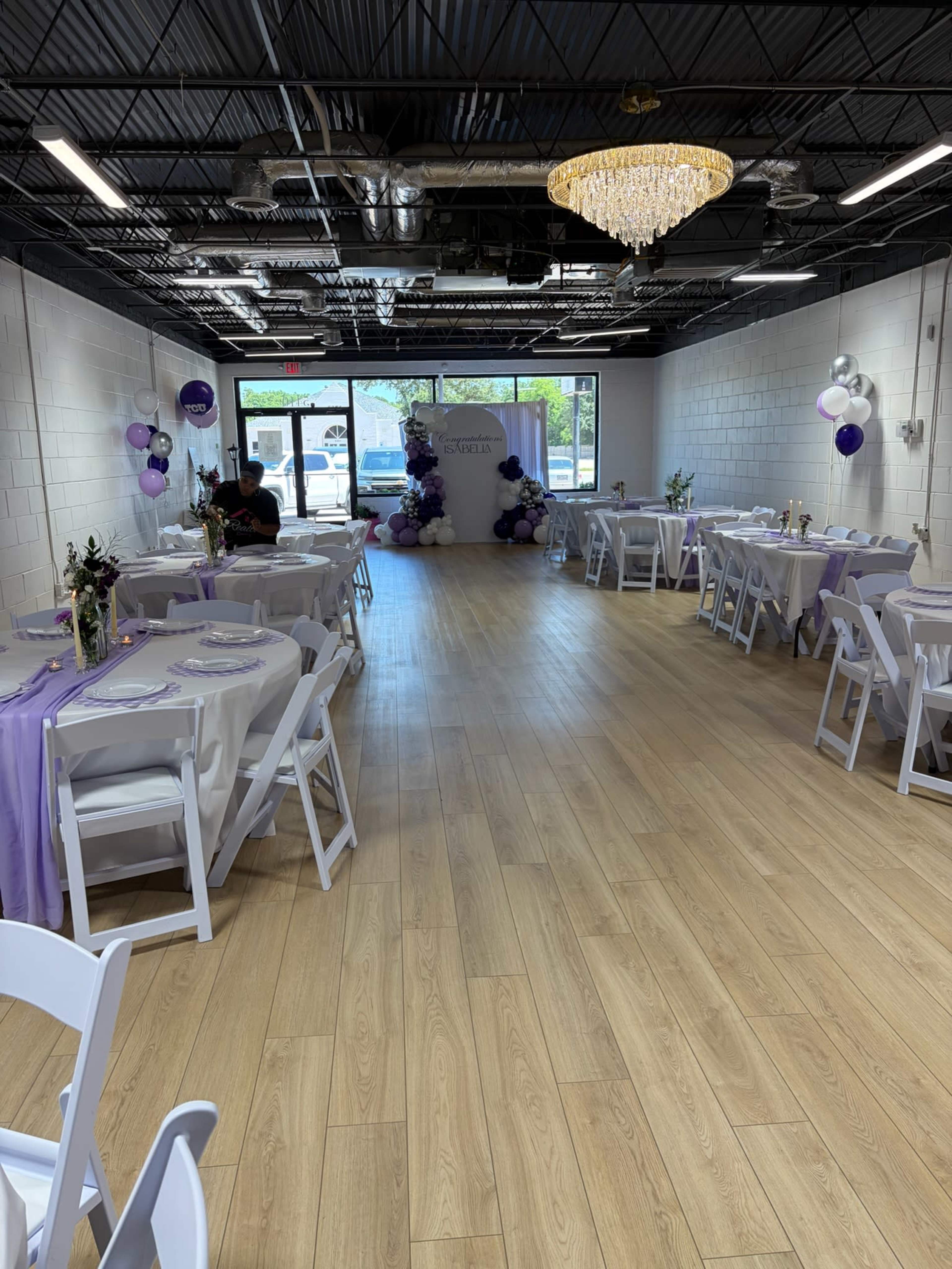 The image shows an indoor event space set up for a gathering, featuring tables dressed in purple and white linens, a decorative backdrop, and balloons in the corners.