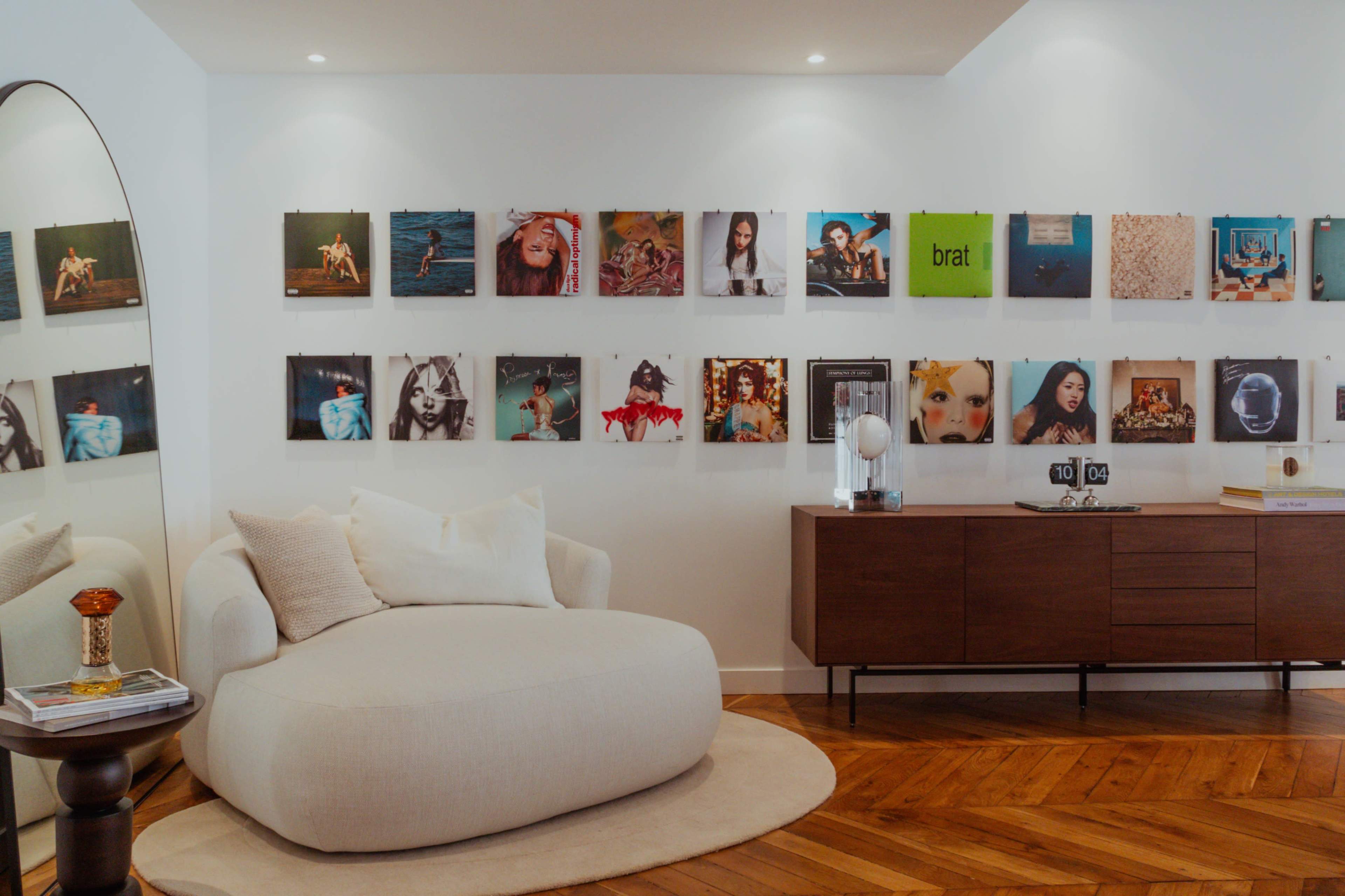 A curved beige sofa sits on a circular rug in a room with a wooden floor, featuring a gallery wall of framed photographs and art.