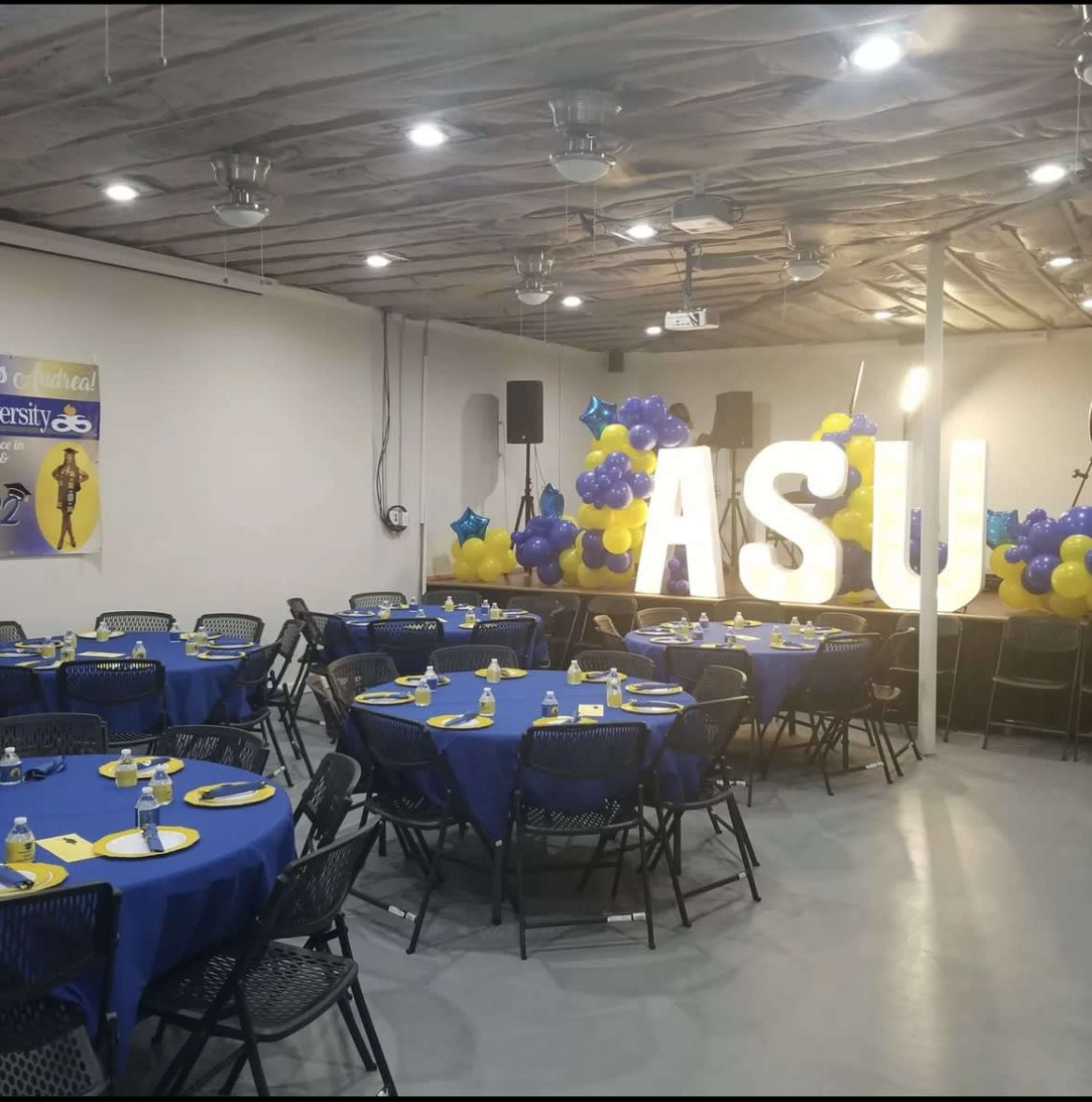 A decorated indoor event space with round tables set with blue tablecloths and yellow accents, featuring large illuminated letters "ASU" and a balloon display in blue and yellow.