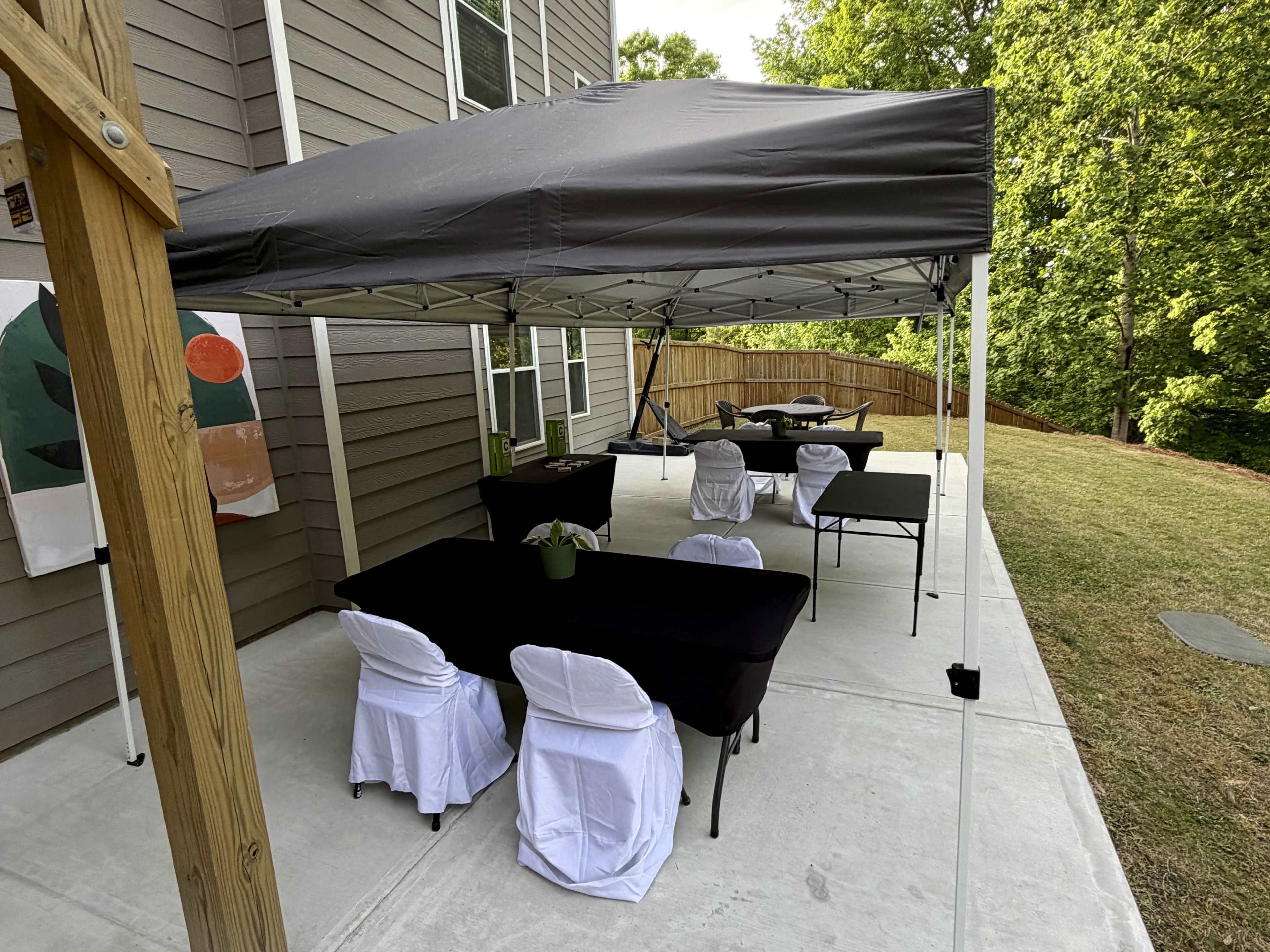 A black canopy is set up over tables with white chair covers in a backyard space next to a house.