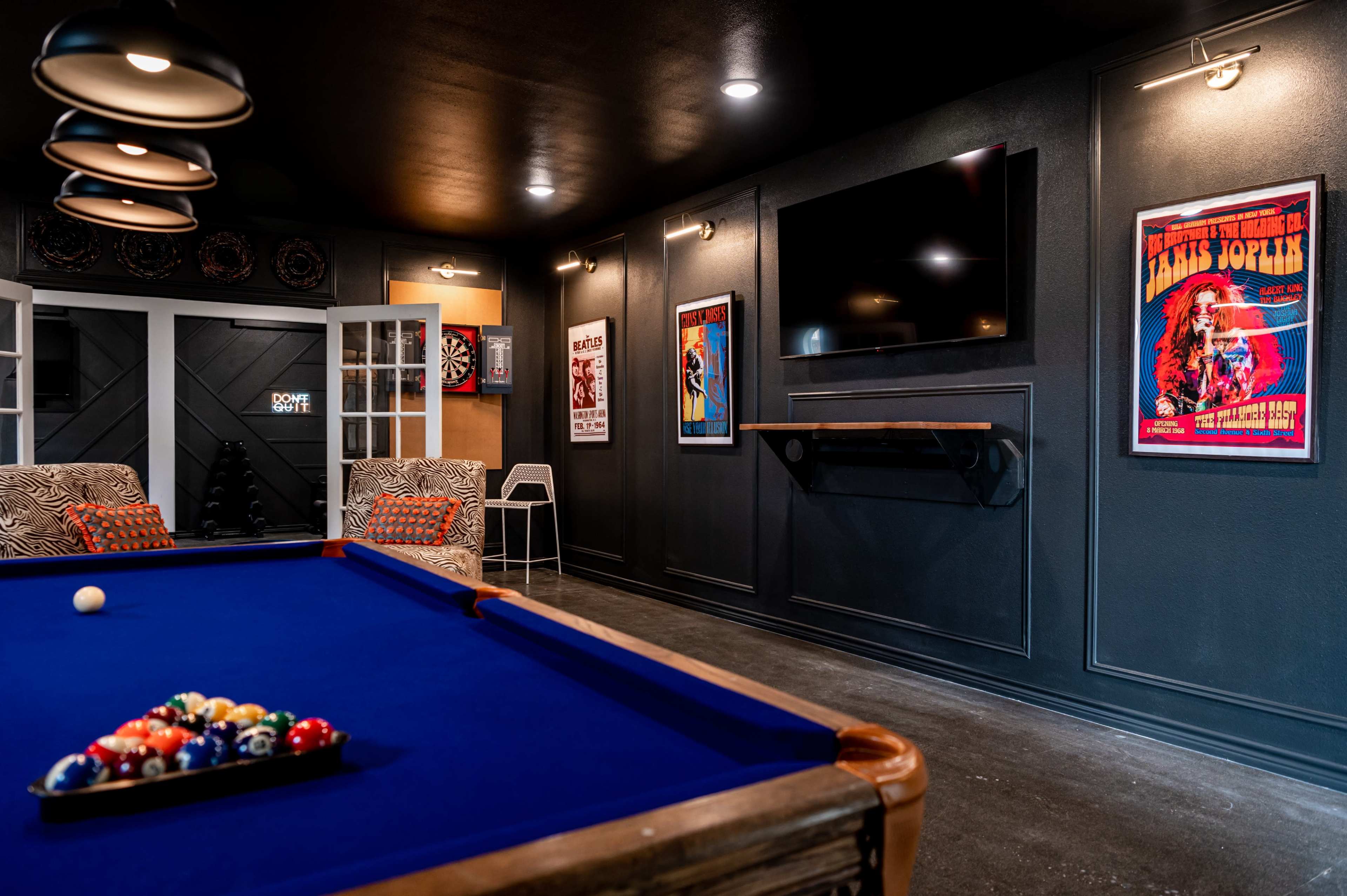 A billiards room features a blue pool table with a triangle of colored balls and framed posters on the dark walls.