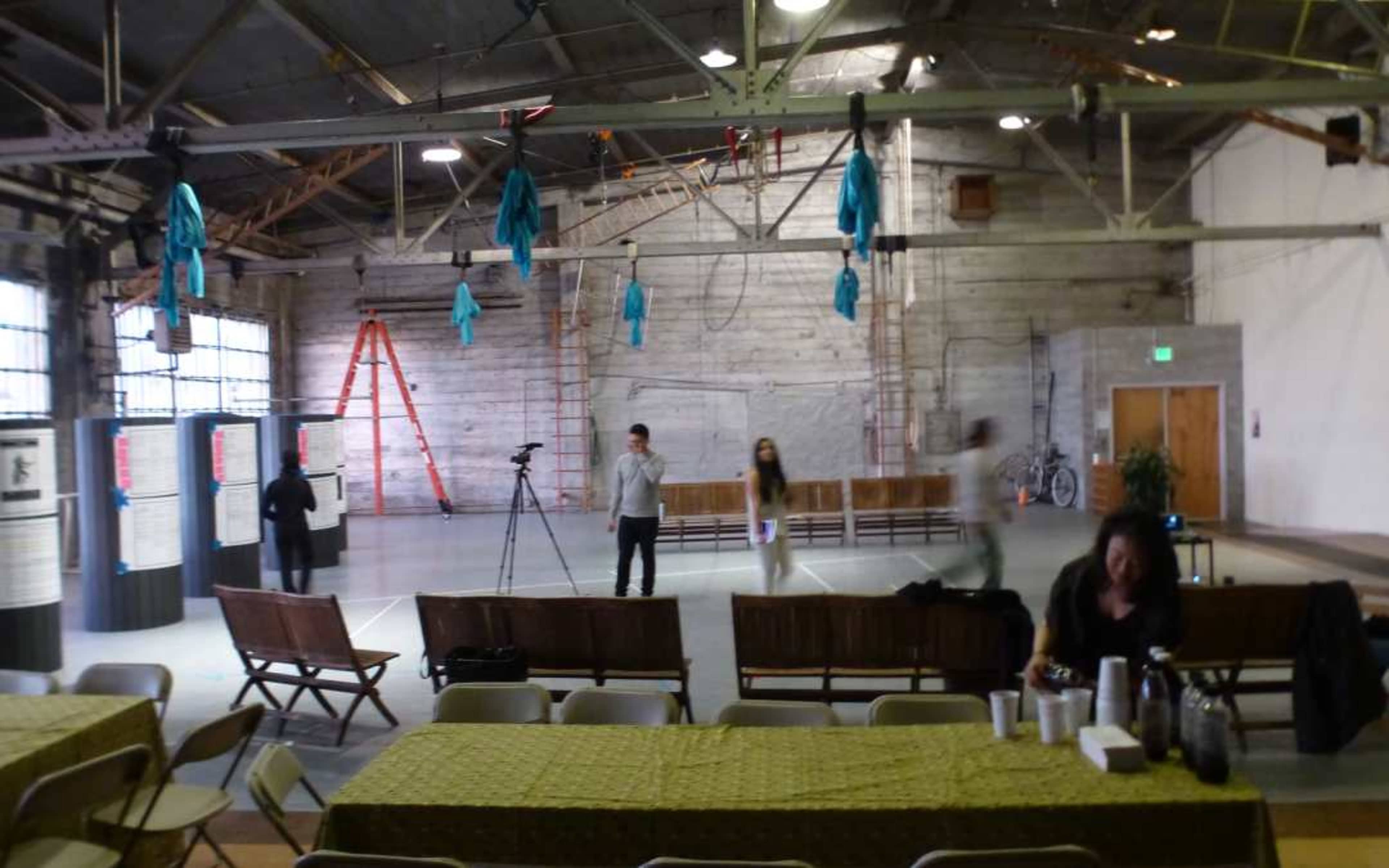 The image shows an industrial space with wooden benches, a few people moving around, and blue cloths hanging from the ceiling.