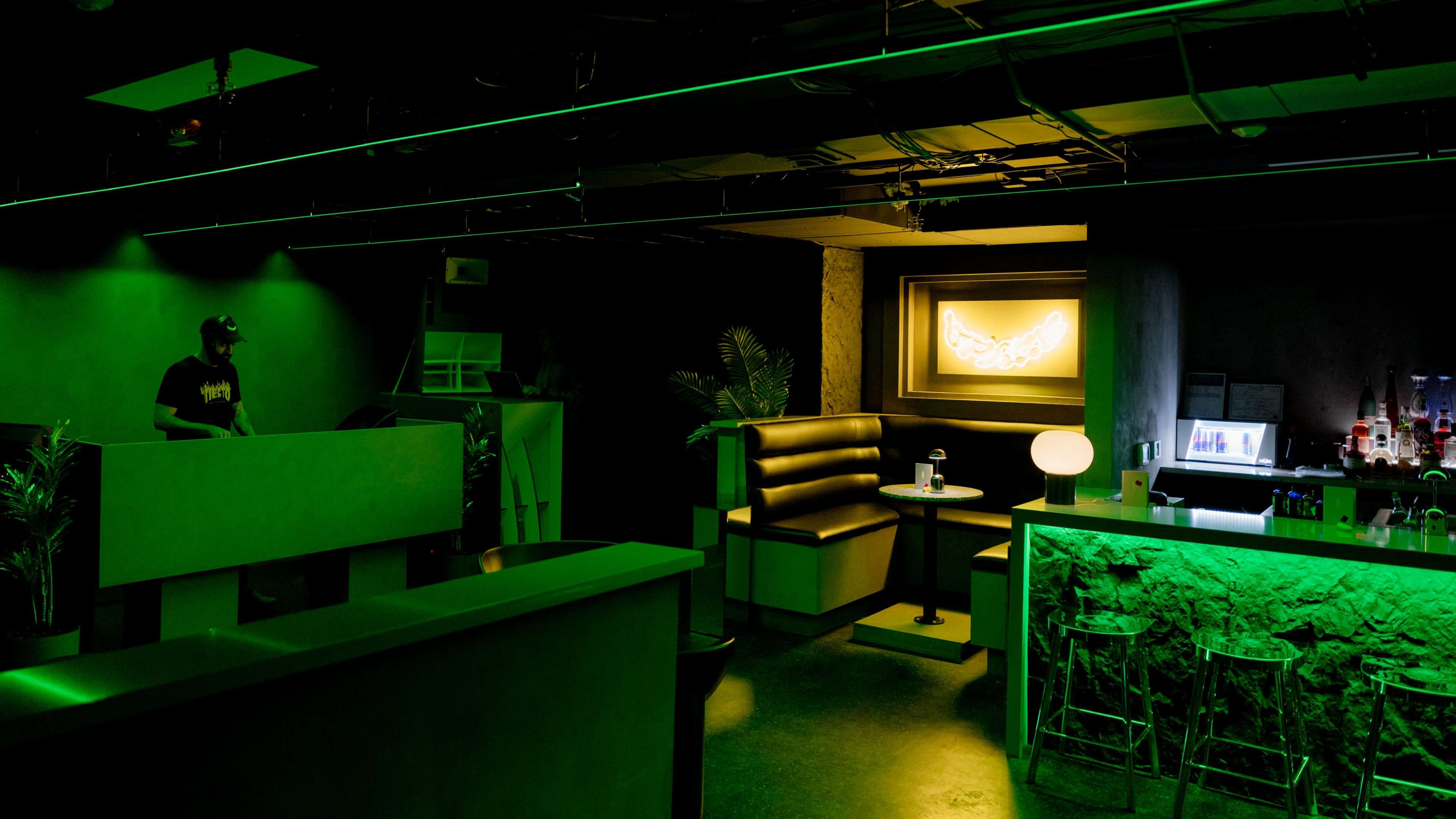 A dimly lit lounge features green lighting, a bar with illuminated decor, and cozy seating areas.