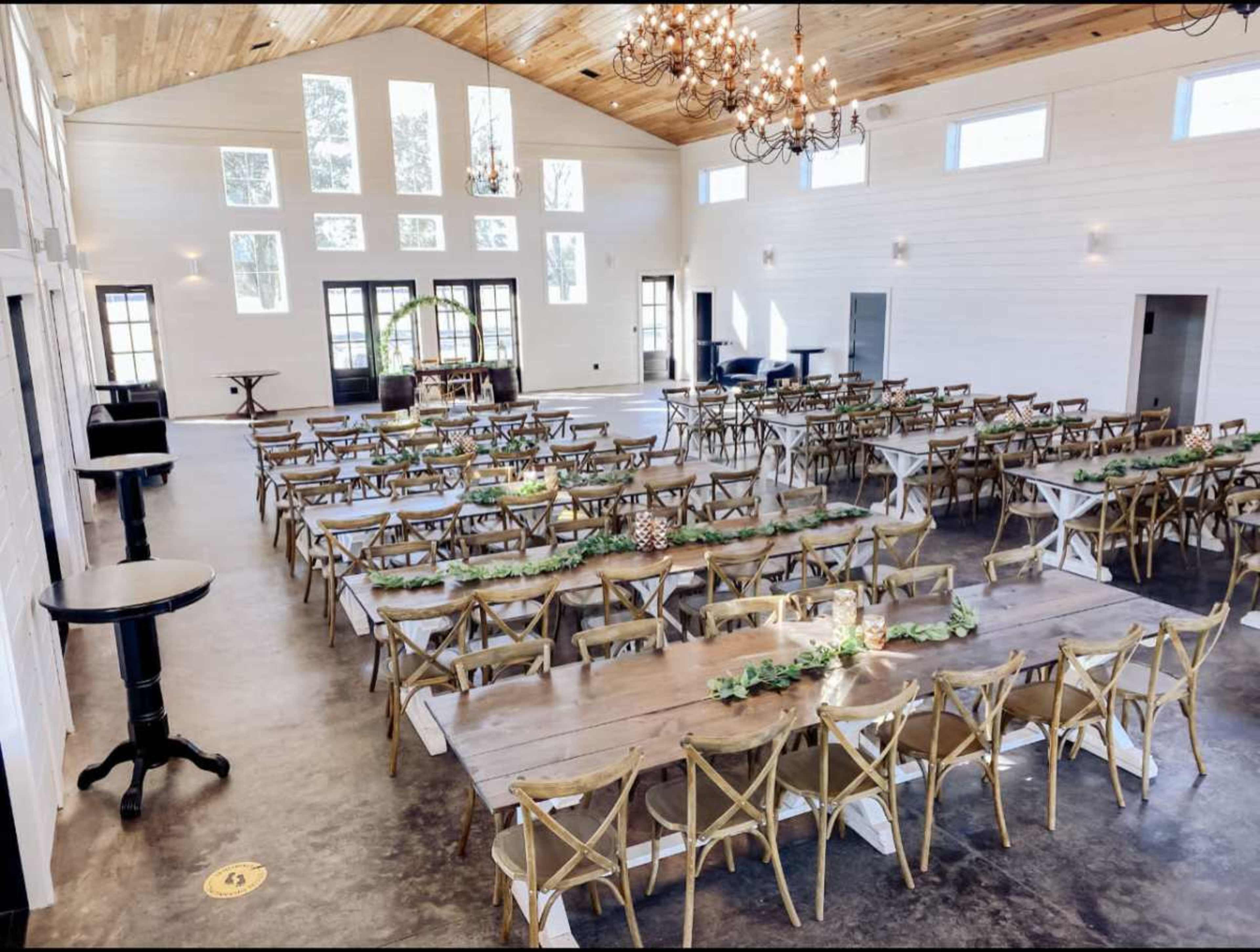 The image shows a large, elegantly arranged banquet hall with wooden tables and chairs set for an event, featuring high ceilings and bright windows.