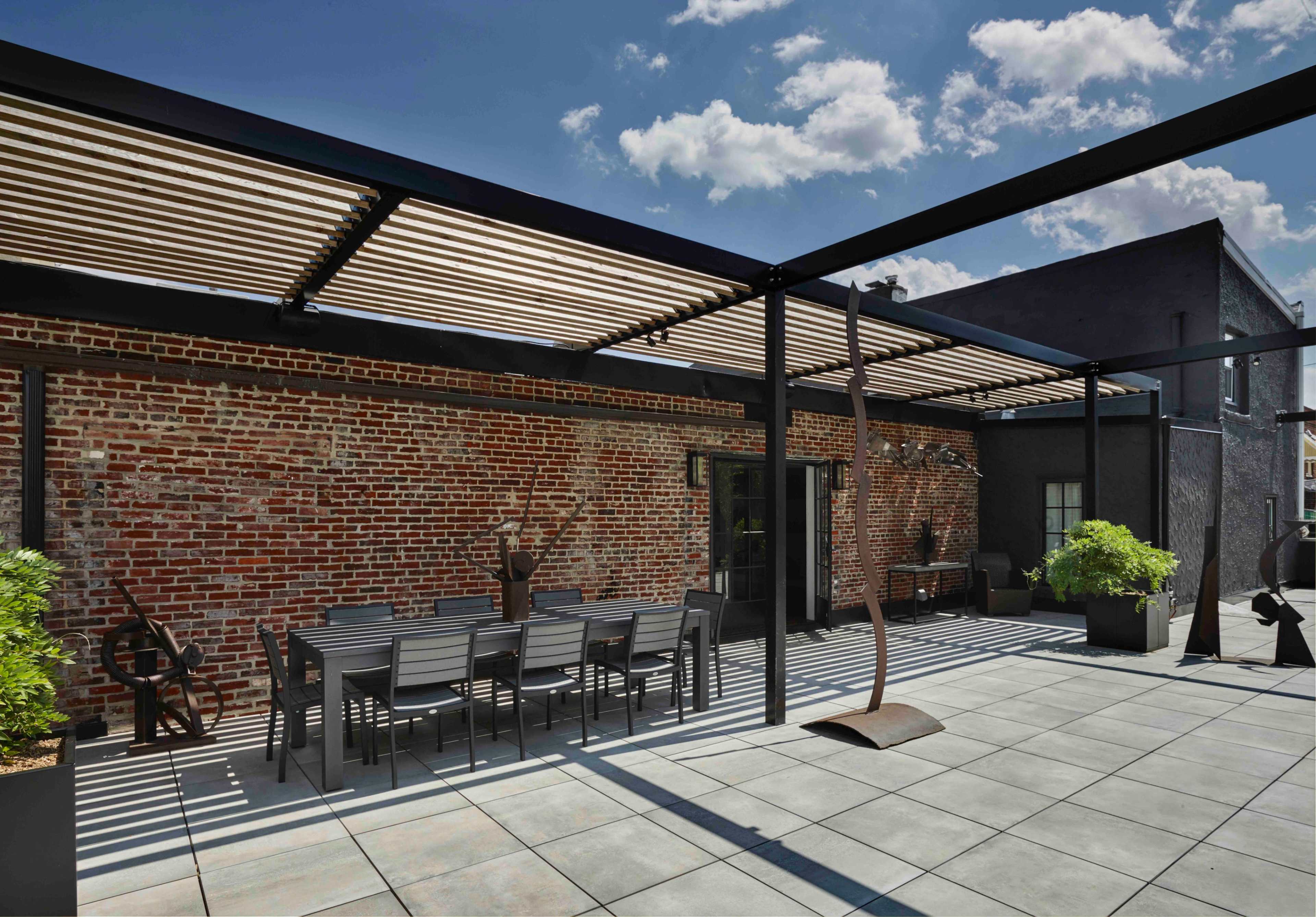 The image shows a spacious outdoor patio with a dining table, surrounded by potted plants and a brick wall, under a modern slatted overhead structure.