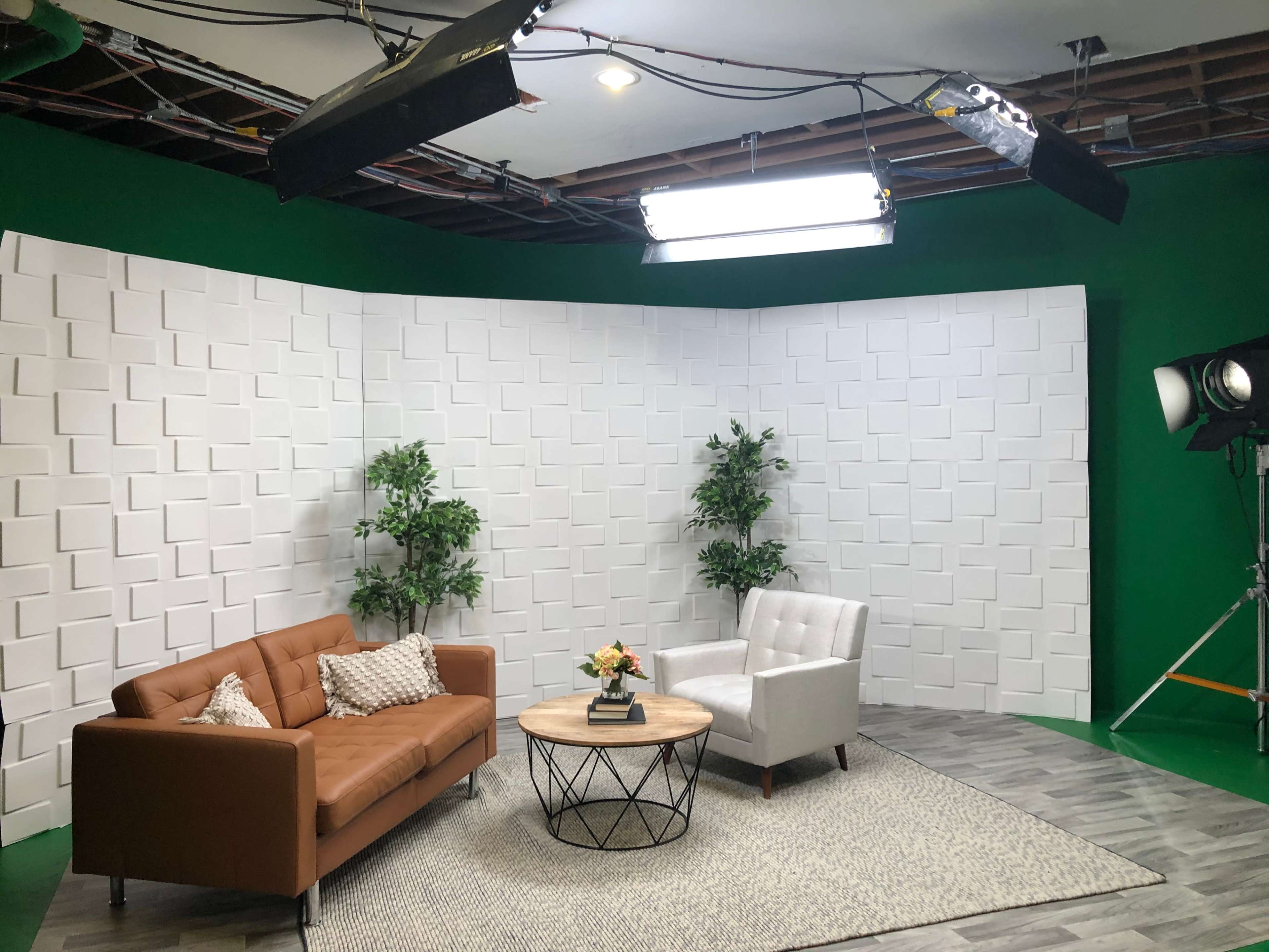 A TV studio set with a brown sofa, a white armchair, a round coffee table, two plants, and a textured white backdrop.