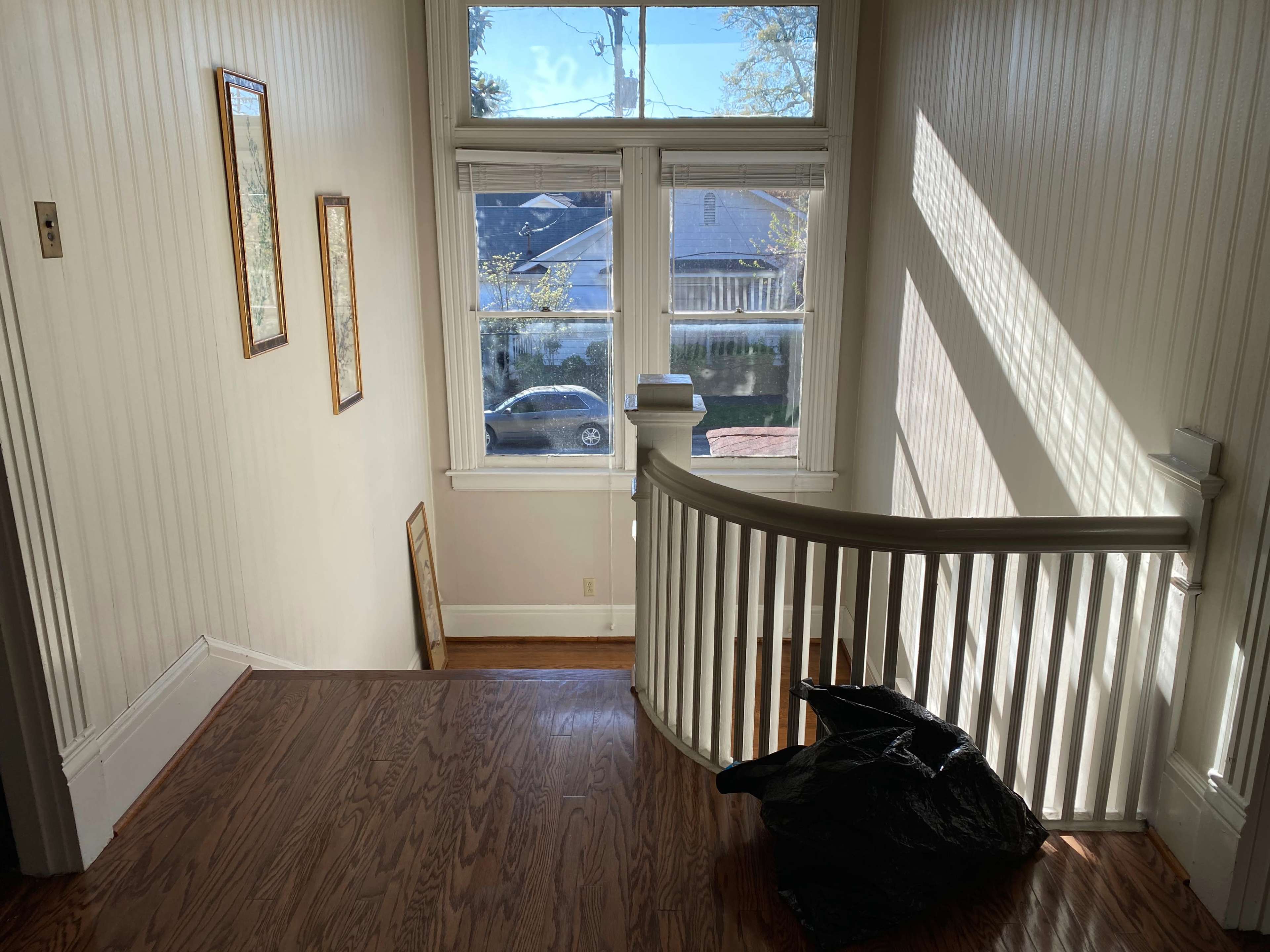 A well-lit staircase leads down to a lower level, with a large window providing natural light and a black garbage bag resting on the wooden floor.