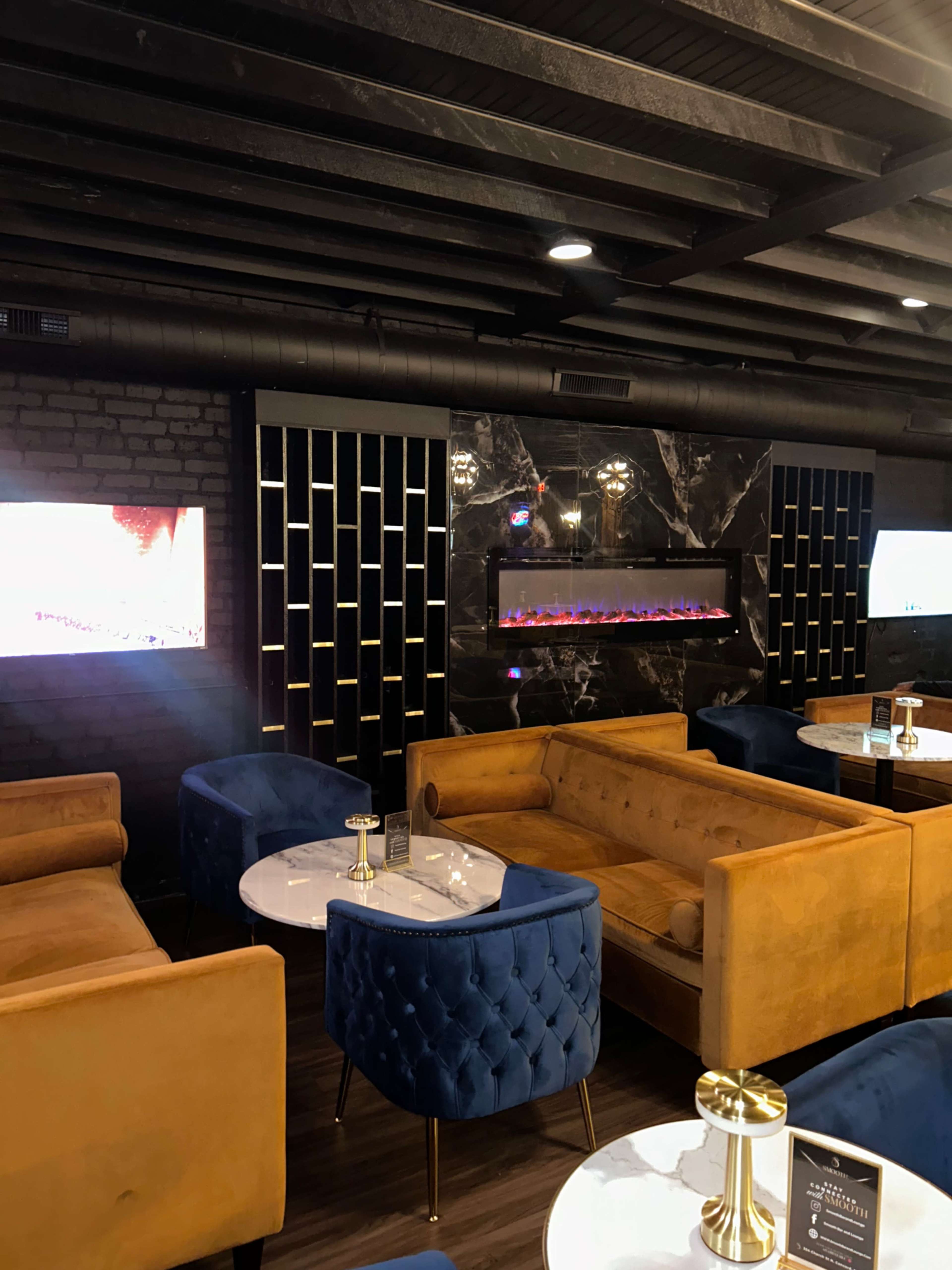 The image shows a modern lounge with yellow and blue upholstered sofas, marble tables, and a sleek black wall featuring a decorative electric fireplace and two screens.