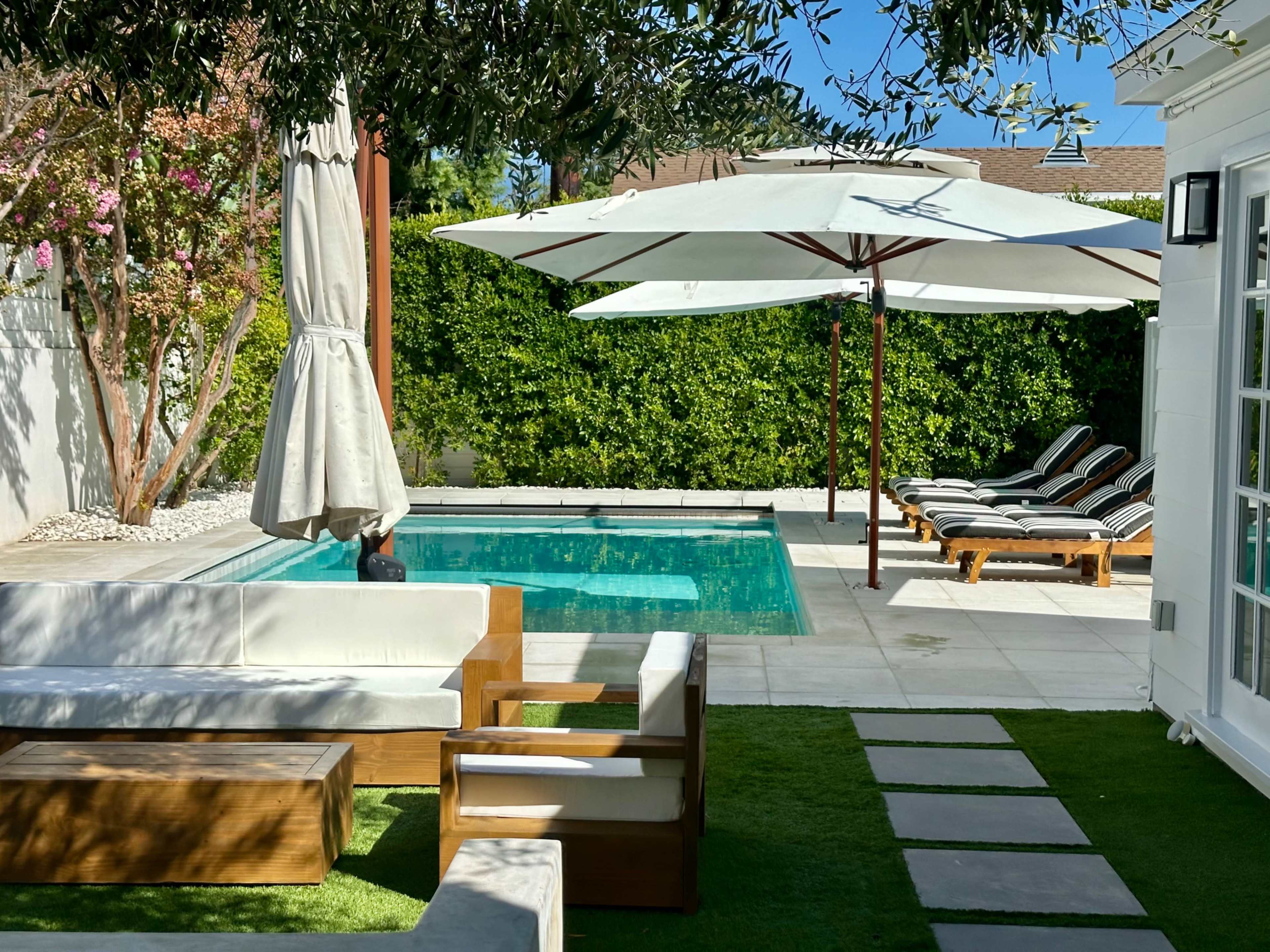 The image shows a tranquil backyard with a swimming pool, lounge chairs, and shaded seating areas surrounded by greenery.