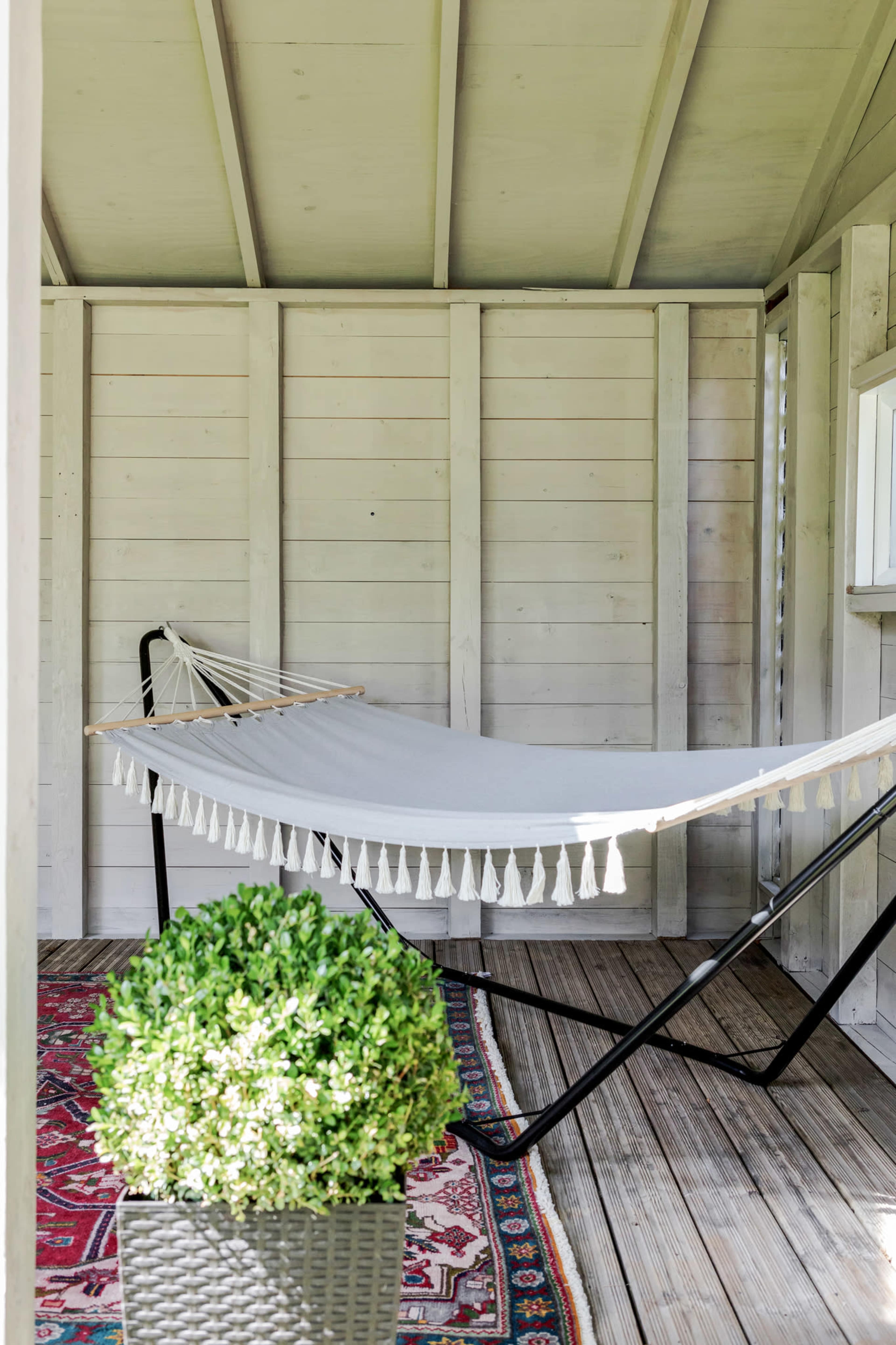A light-colored wooden space with a hammock hung on a black frame and a potted green shrub in the foreground.