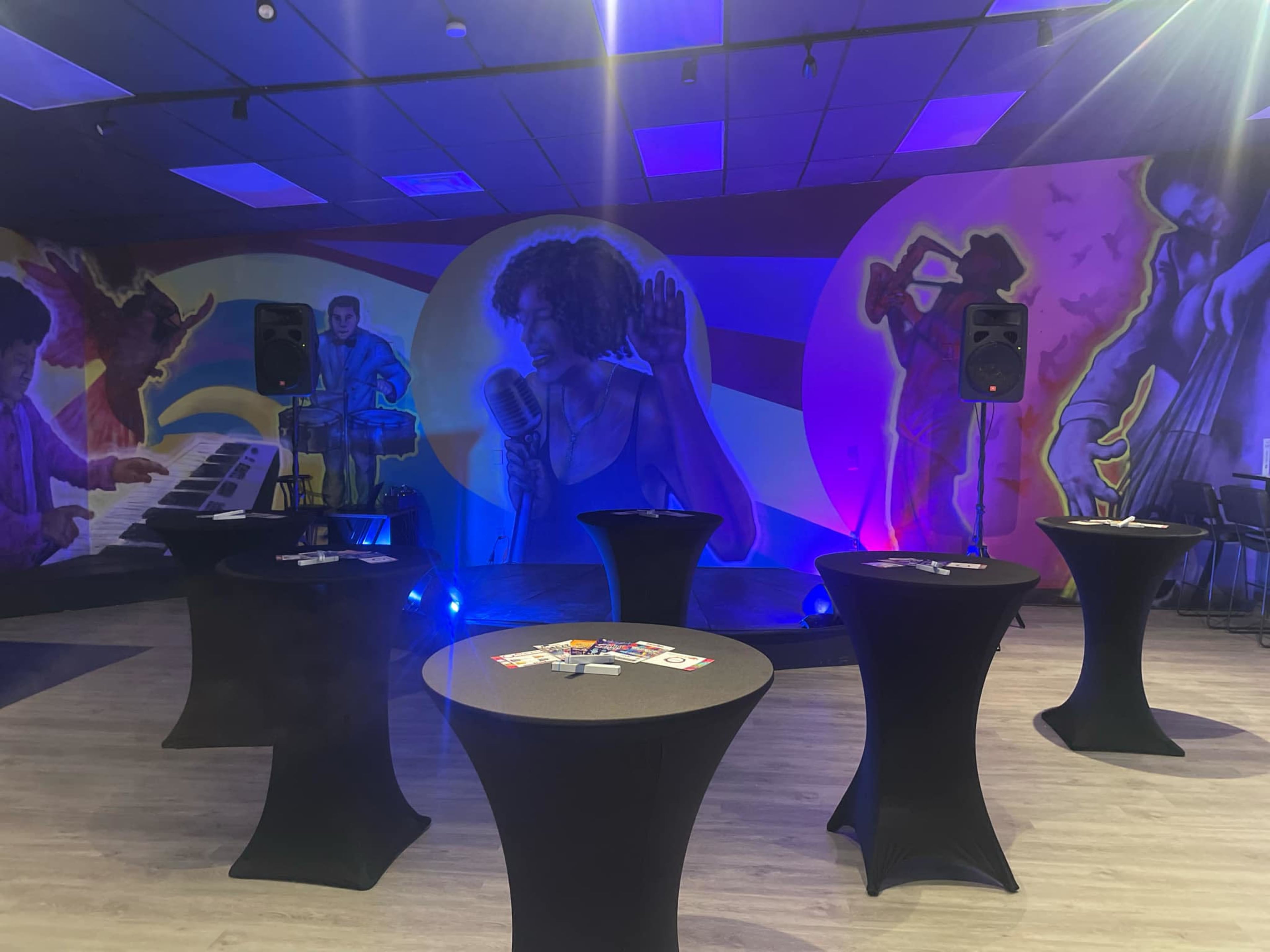 The image shows a vibrant event space featuring round black tables, a DJ setup, and a large mural depicting musicians on the walls, illuminated by colorful lighting.