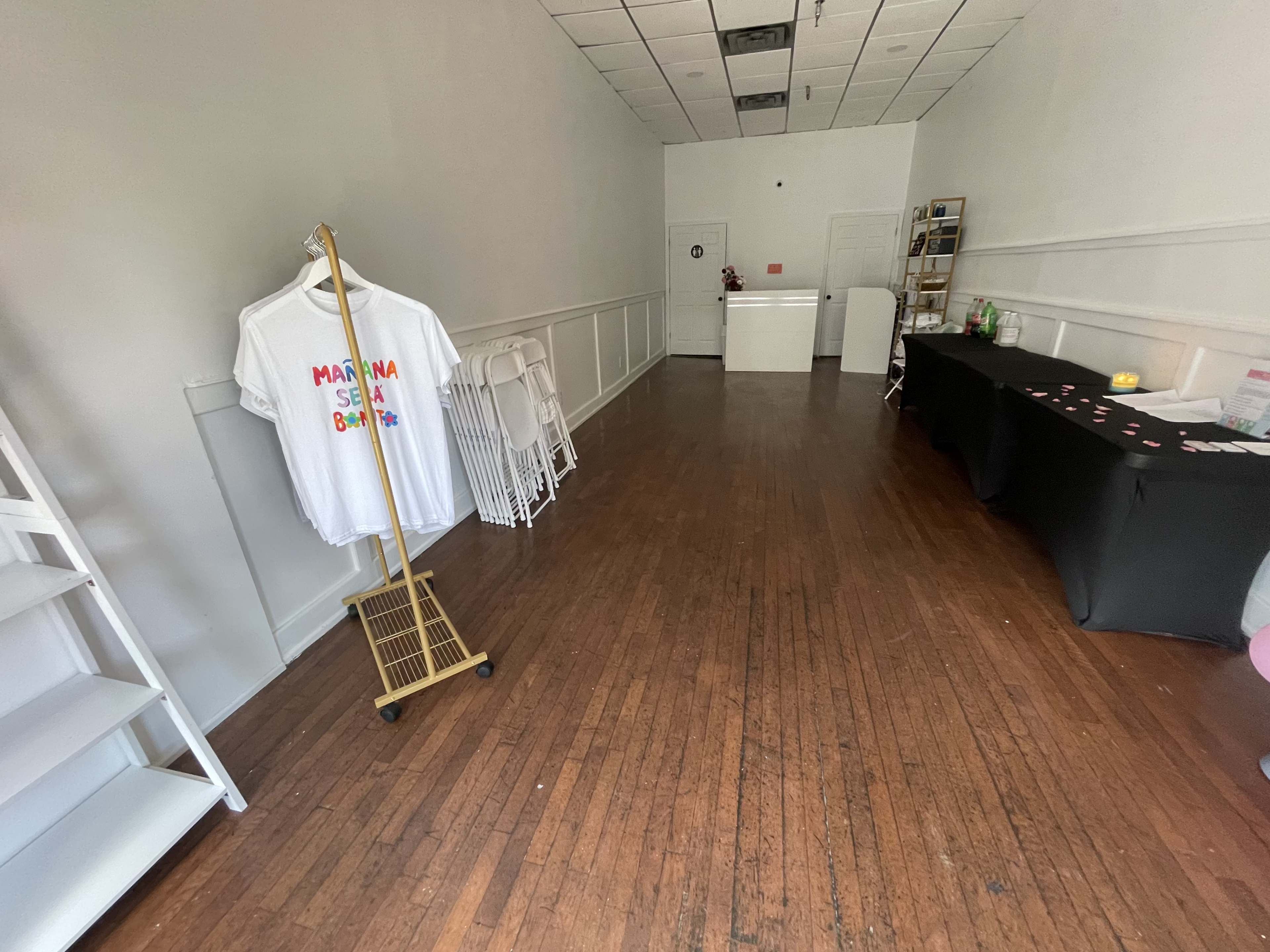 A vacant store interior with light wood flooring, a reception area at the back, and a clothing rack displaying a t-shirt on the left side.
