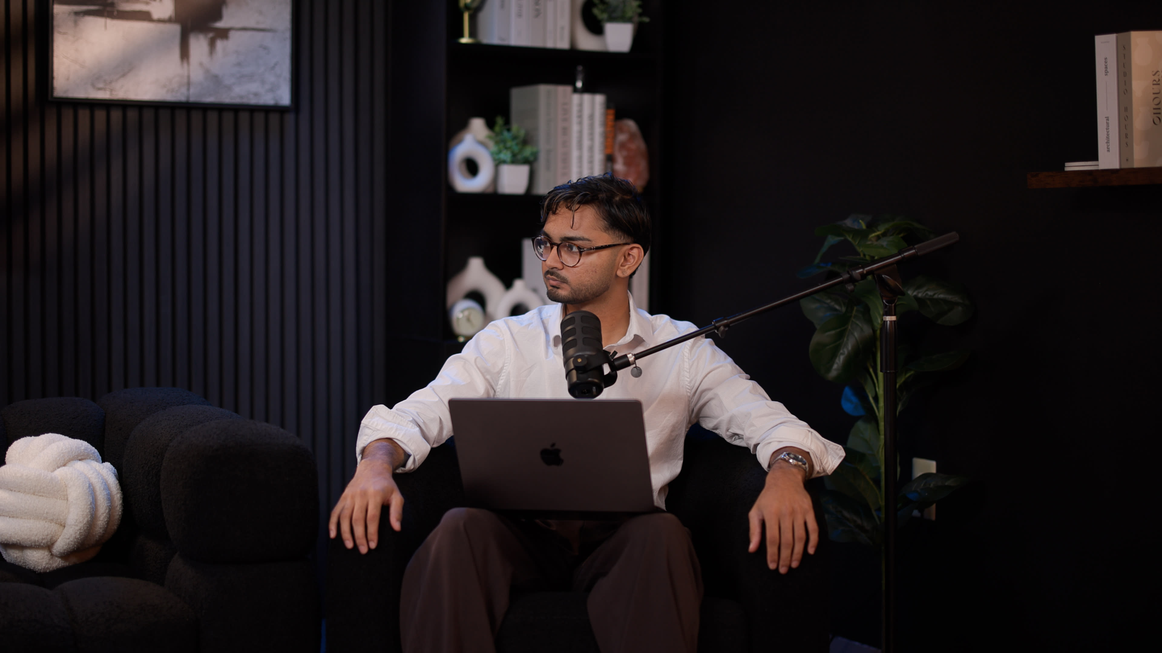 A person sits on a black armchair with a laptop on their lap, facing slightly left, while a microphone is positioned nearby.