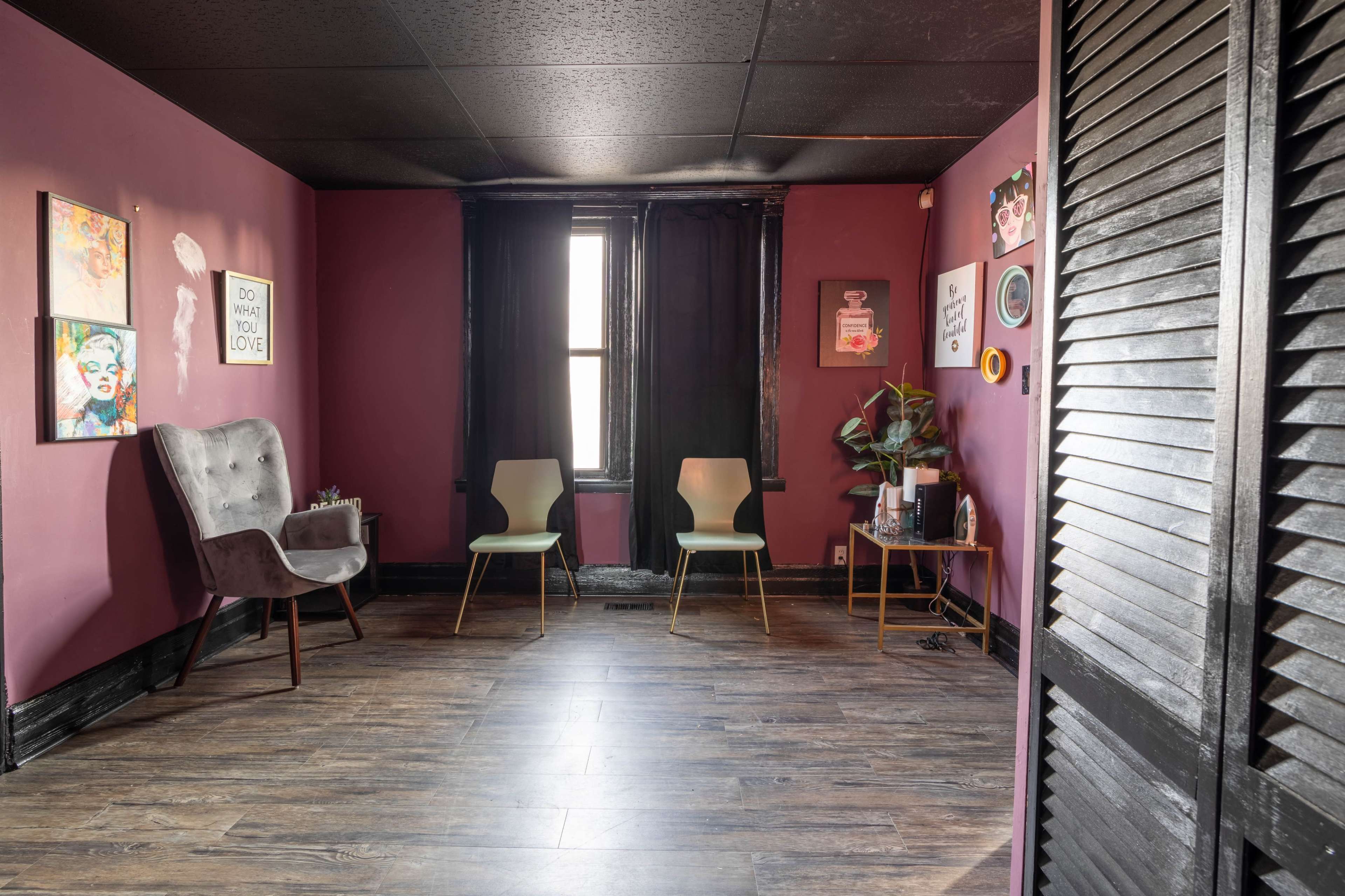 A small, purple-walled room with two chairs and an armchair facing a window, illuminated by natural light.