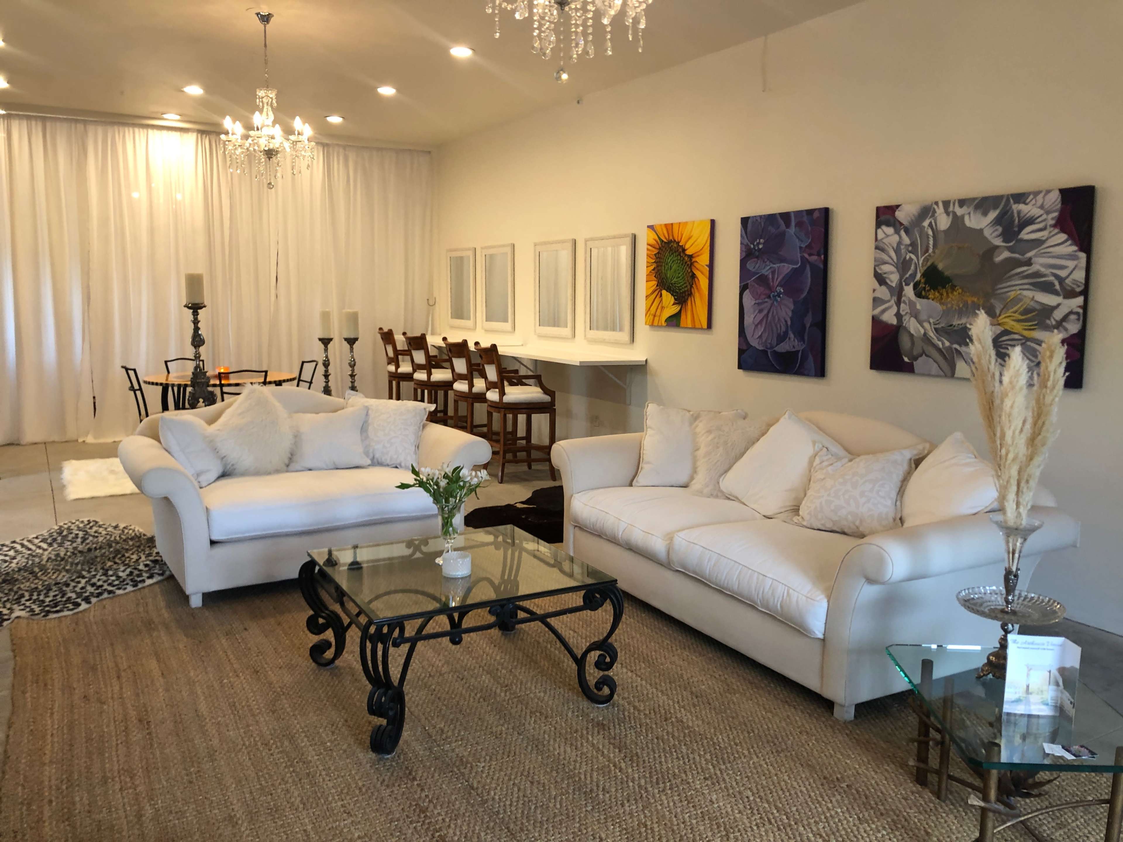A modern living room with white sofas, a glass coffee table, and colorful artwork on the walls, leading into a dining area with a table and chairs.