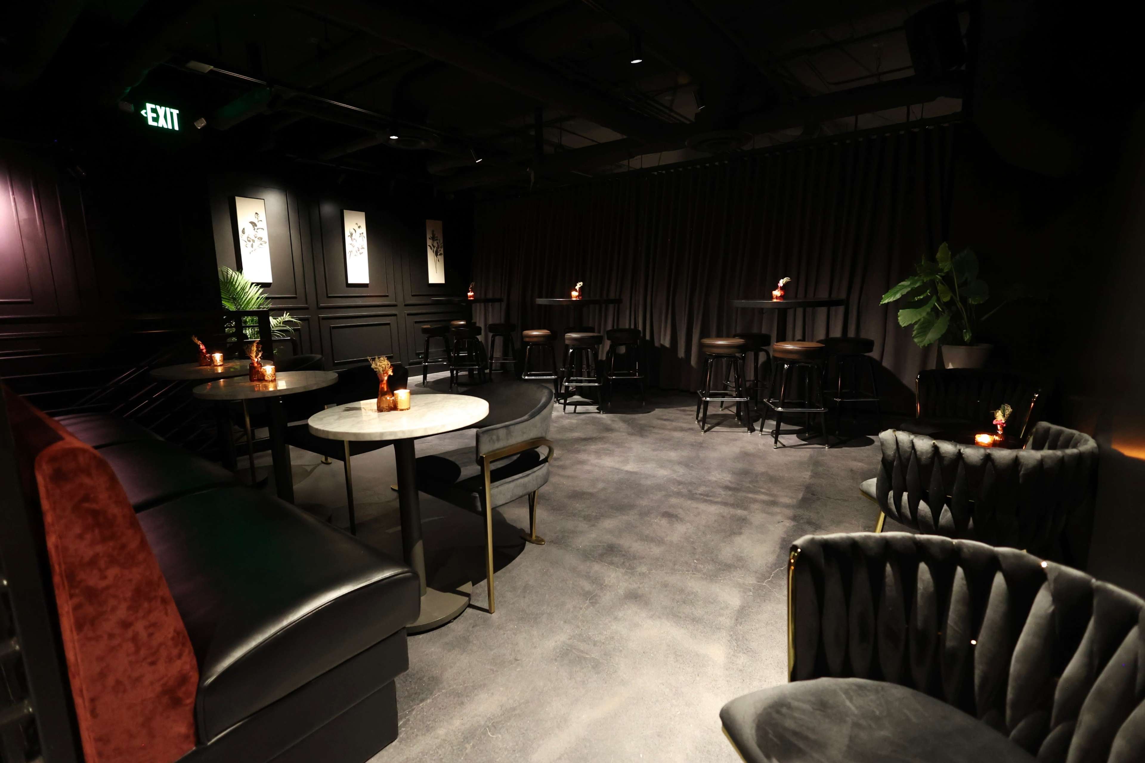 The image shows a dimly lit lounge with a dark color scheme, featuring several tables, chairs, and a few plants against a backdrop of curtains and framed artwork.