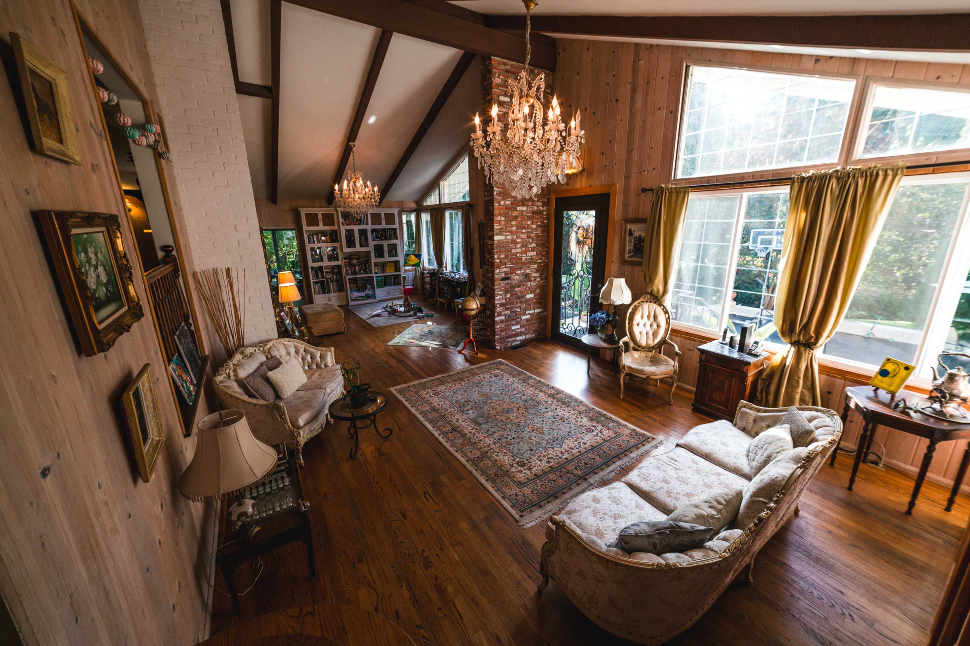 The image shows a spacious living room featuring wooden flooring, large windows with natural light, elegant furniture, and a chandelier.