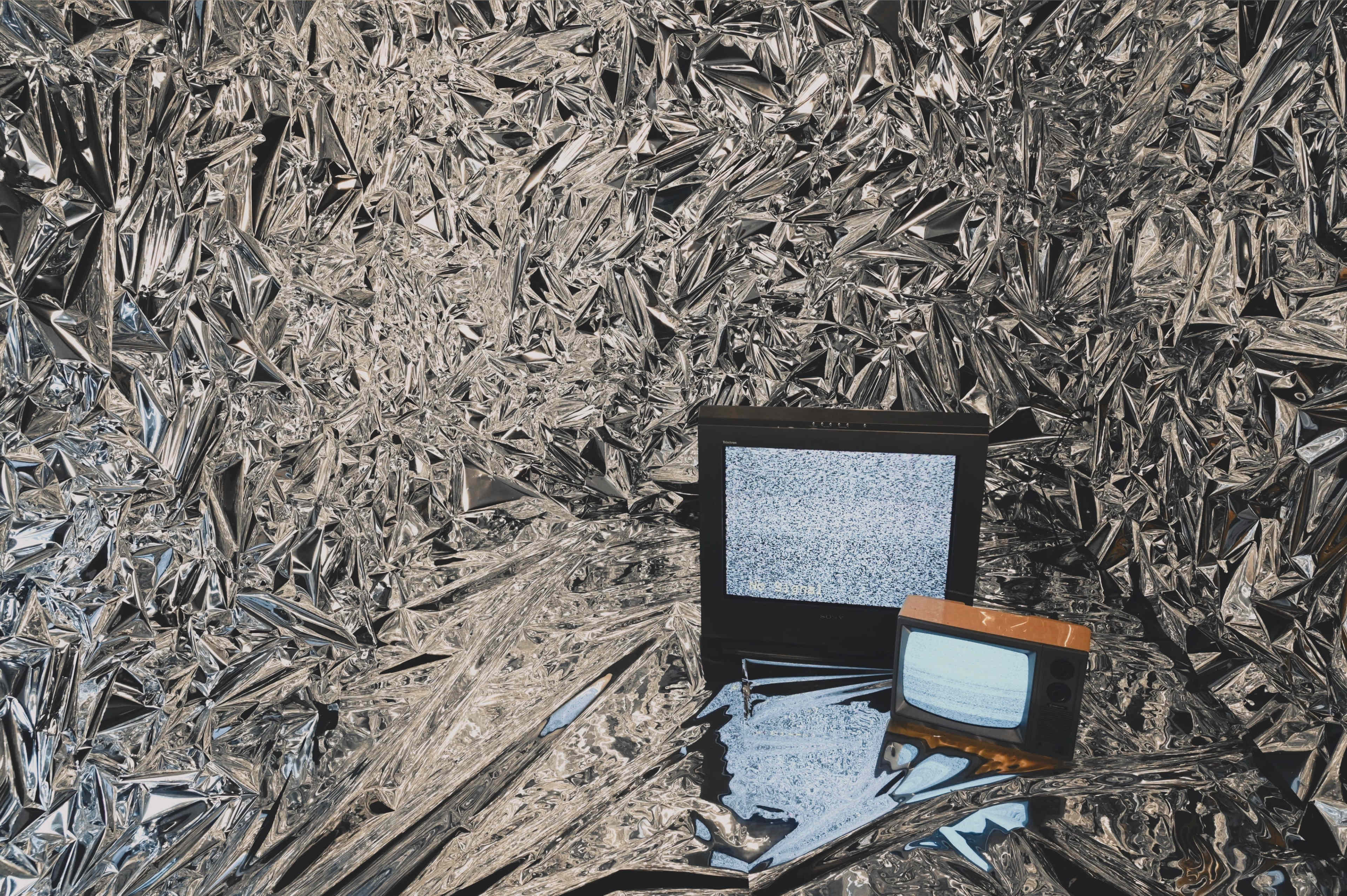 The image shows two old television sets placed on a reflective, foil-covered surface surrounded by shiny, crumpled metallic material.