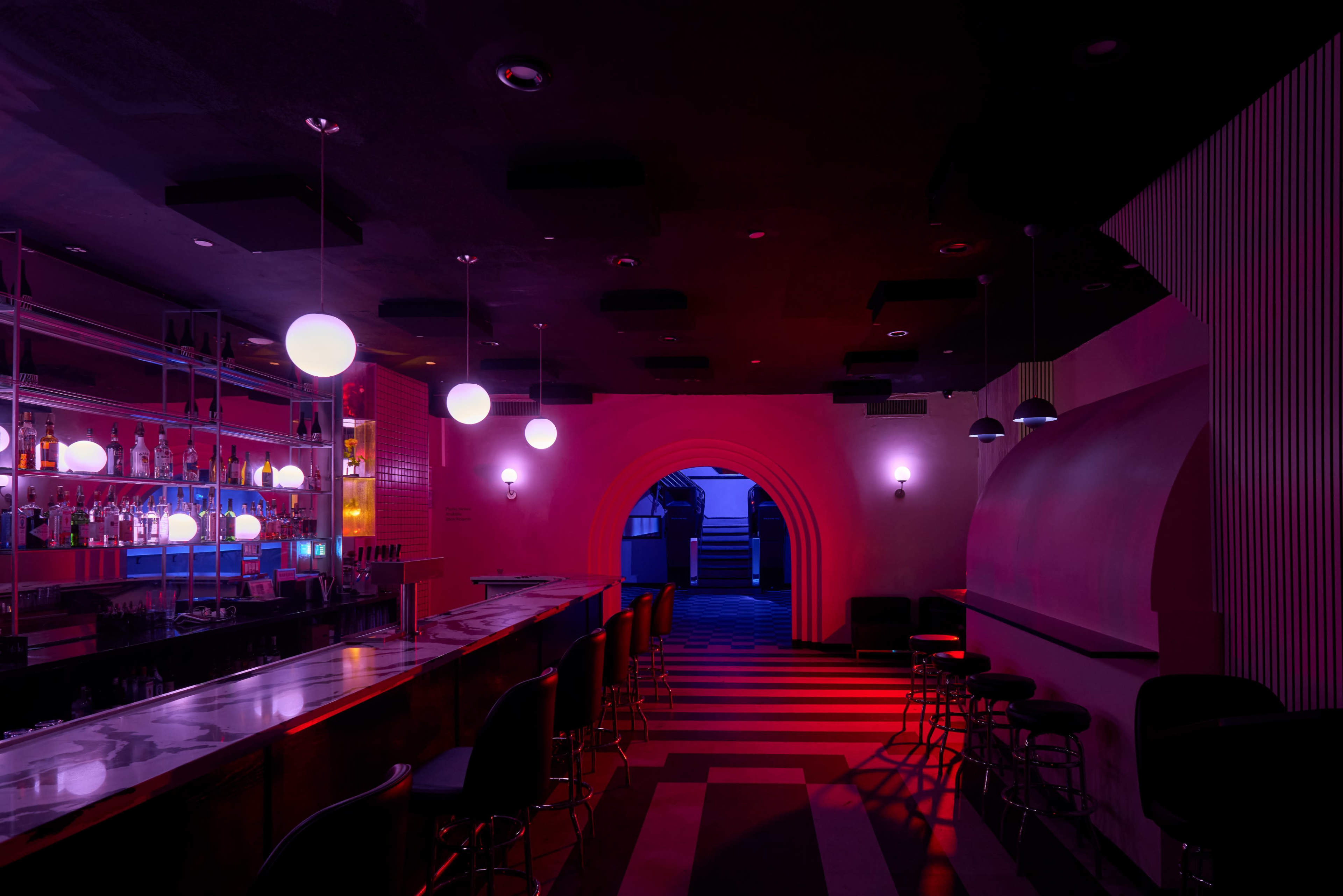 A dimly lit bar area with a long counter, illuminated by hanging round lights, and an archway leading to another space.