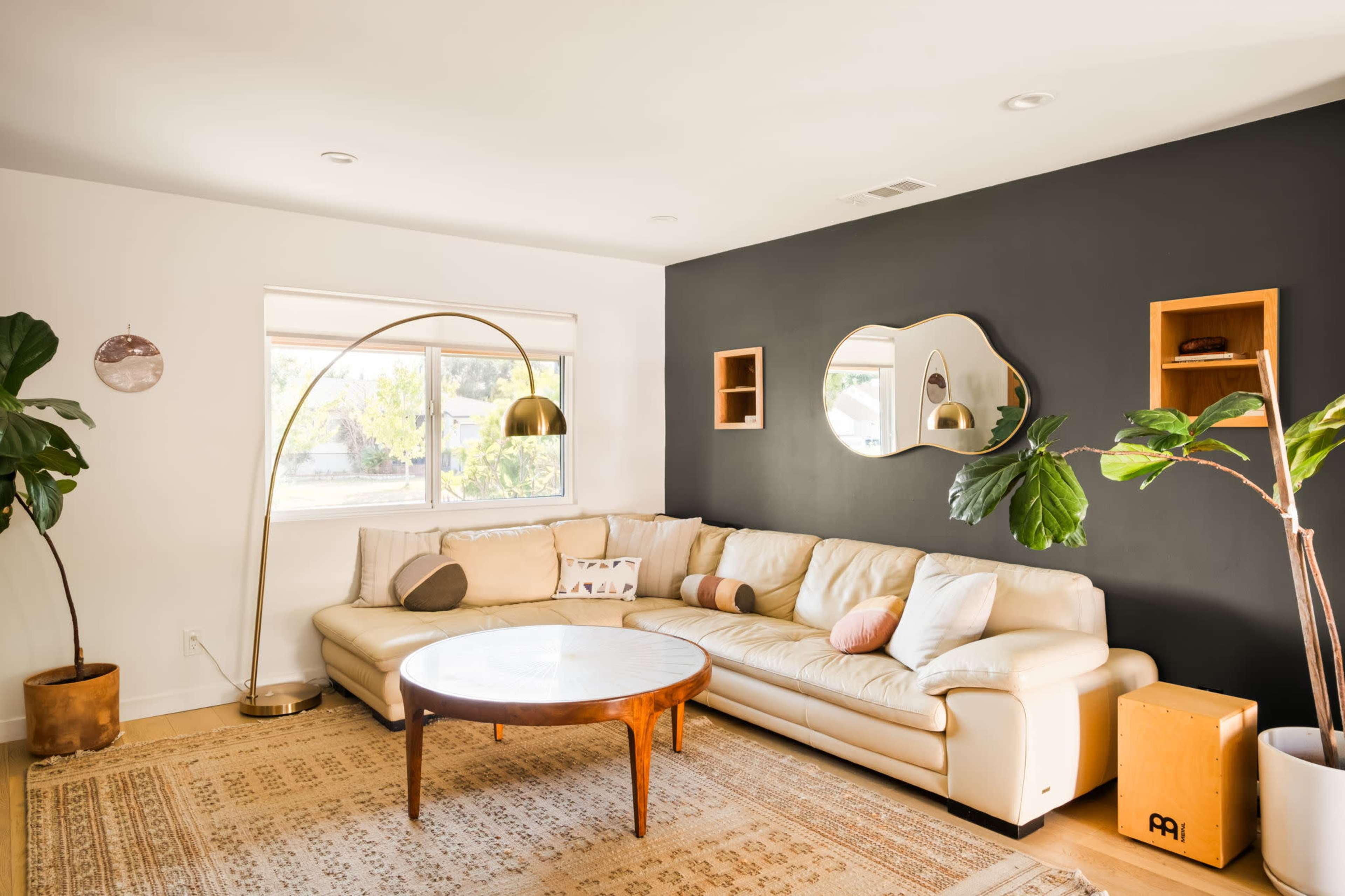 The living room features a beige sectional sofa, a round coffee table, a large floor lamp, and a potted plant, complemented by a dark accent wall and wooden shelves.