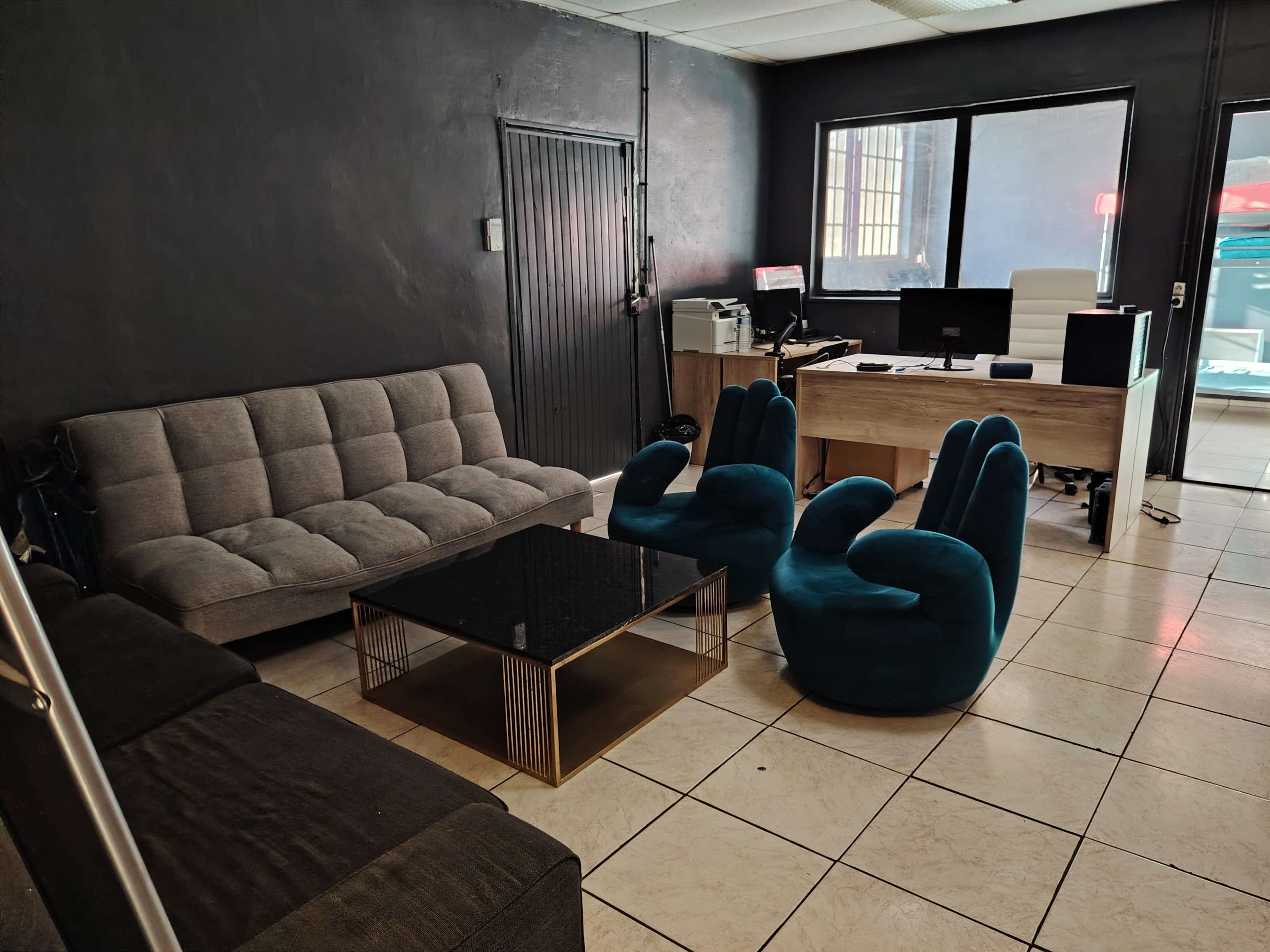The image shows a modern office space with a gray sofa, a glass coffee table, two blue lounge chairs, and a wooden desk, all set against dark walls.