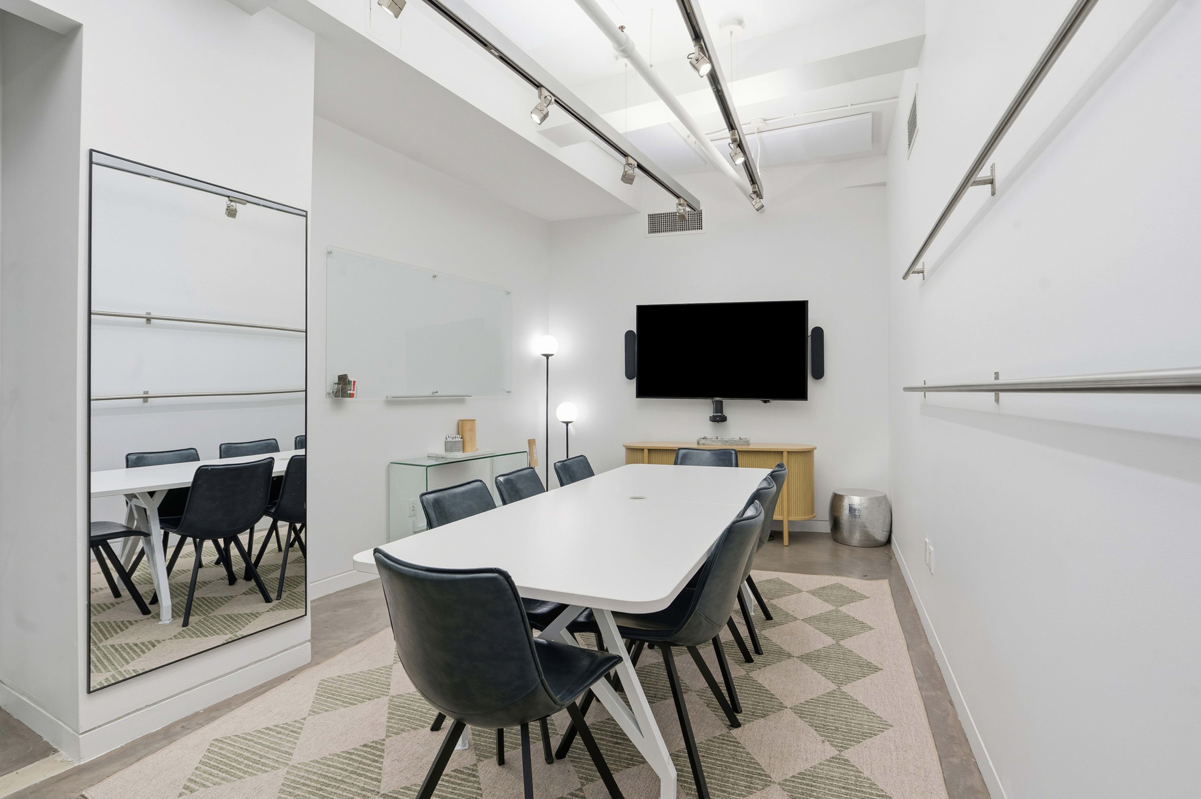 A modern meeting room features a rectangular table surrounded by chairs, a television mounted on the wall, a mirror, and minimalistic decor.