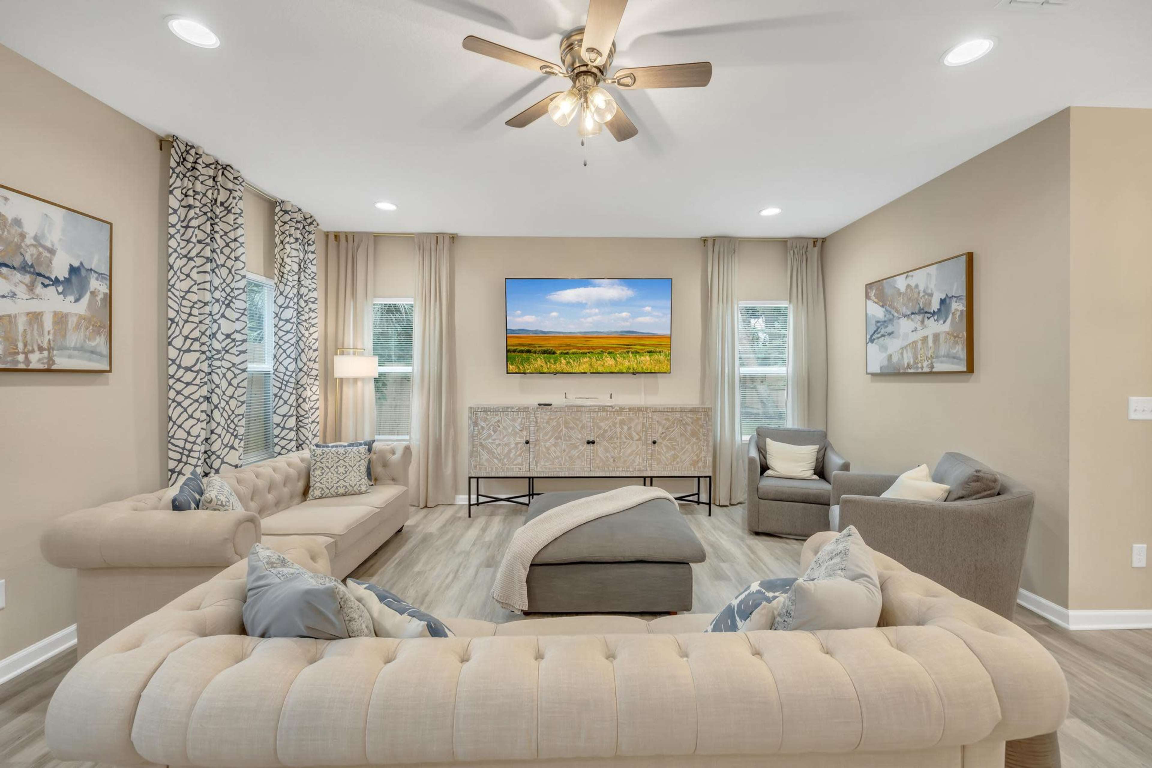 The living room features a light-colored sectional sofa, a gray armchair, a large TV mounted on the wall, and decorative art pieces, all arranged around a central coffee table.
