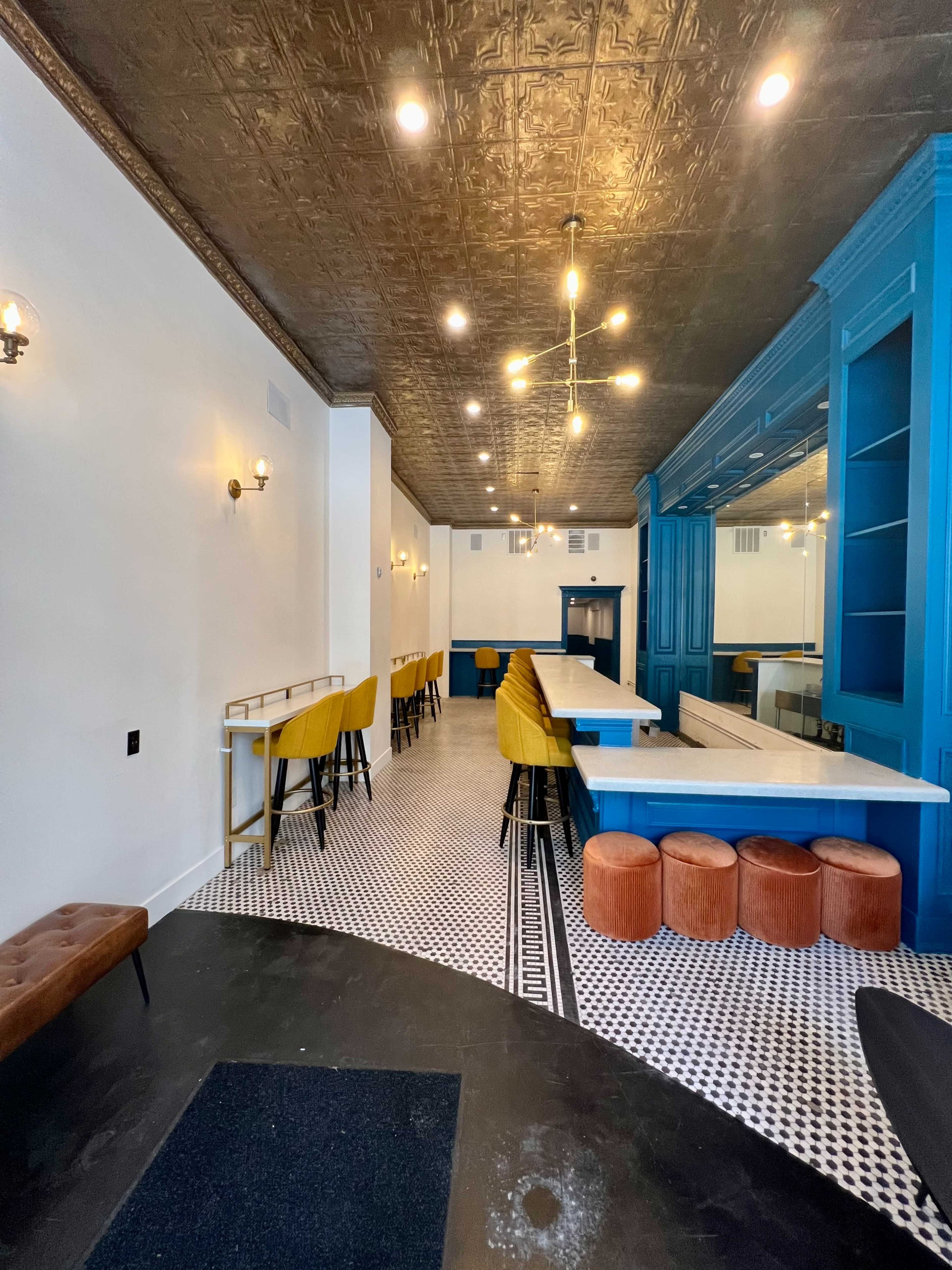 A modern interior of a café or bar featuring a long white bar counter with yellow and brown seating along patterned black and white tile flooring.
