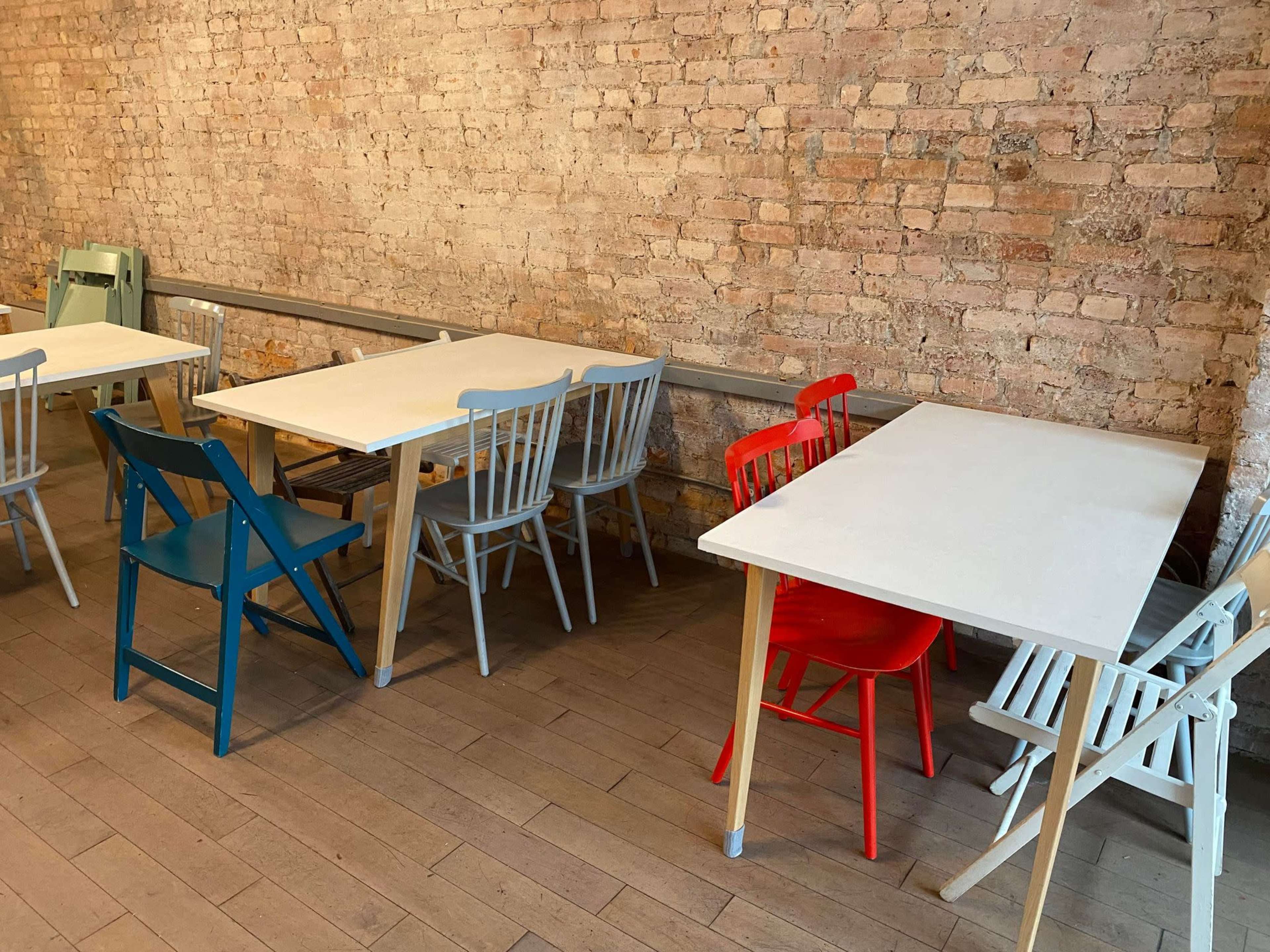 The image shows several tables and chairs in a room with a brick wall, featuring a mix of colored wooden furniture.