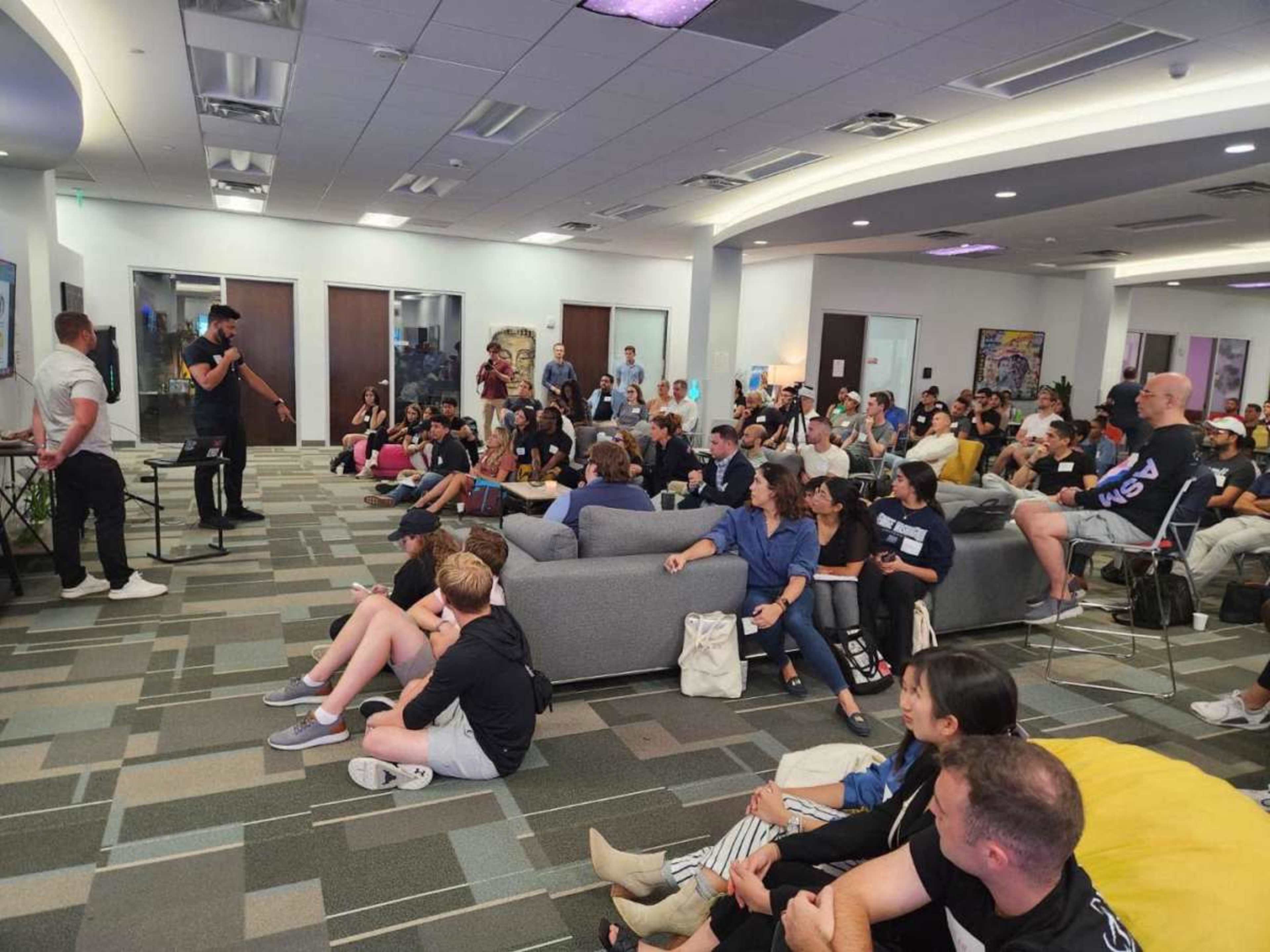 A group of people is seated in a casual setting, listening to a speaker who is standing on a stage in front of them.