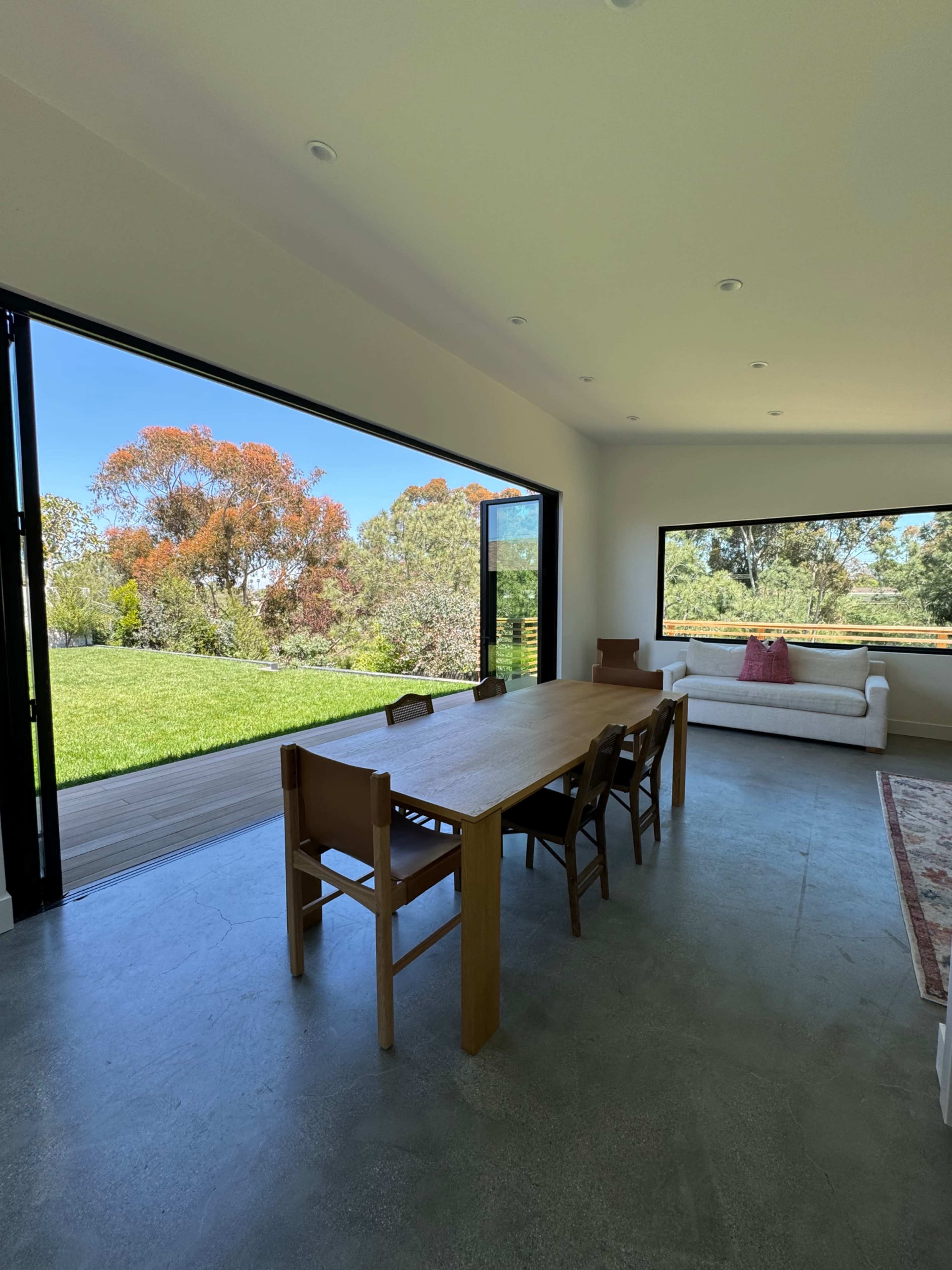 A spacious interior features a wooden dining table with chairs, leading to large sliding glass doors that open to a green landscape.