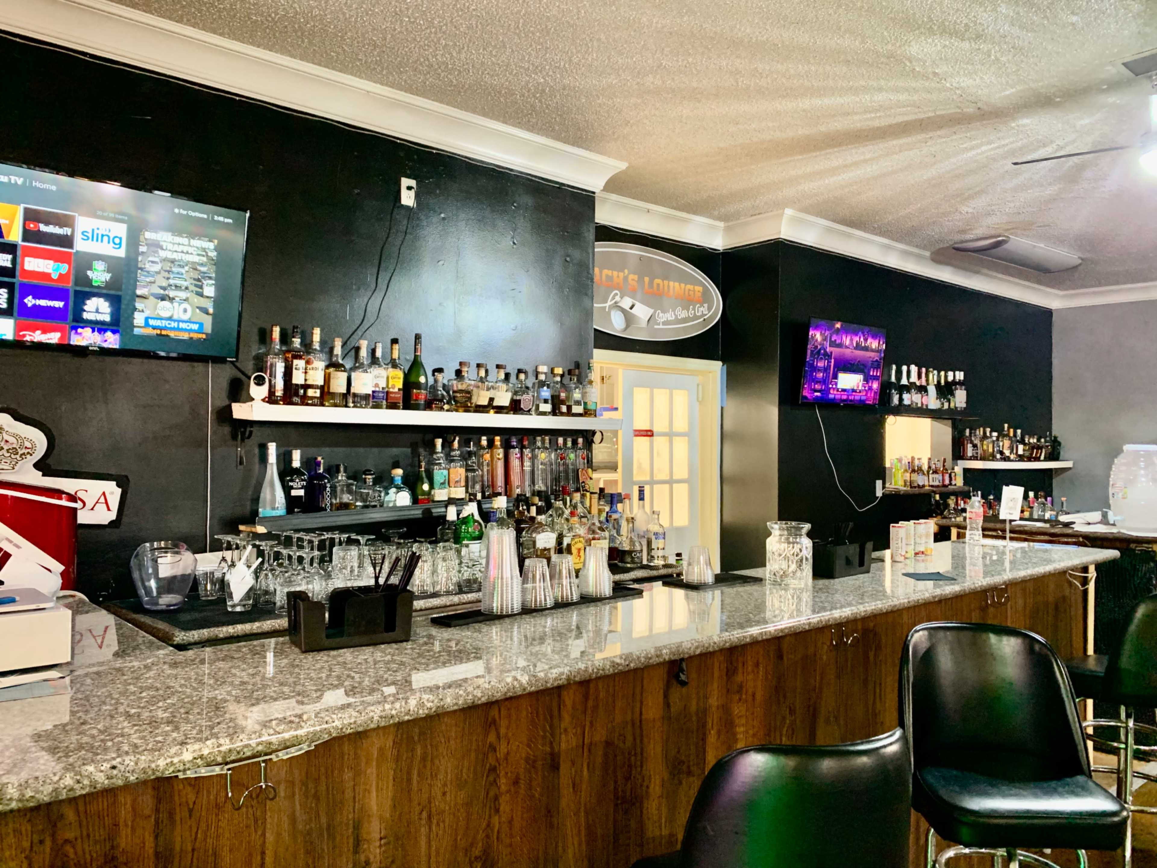 A bar with a sleek granite countertop, shelves lined with various bottles, and two wall-mounted TVs displaying menus.