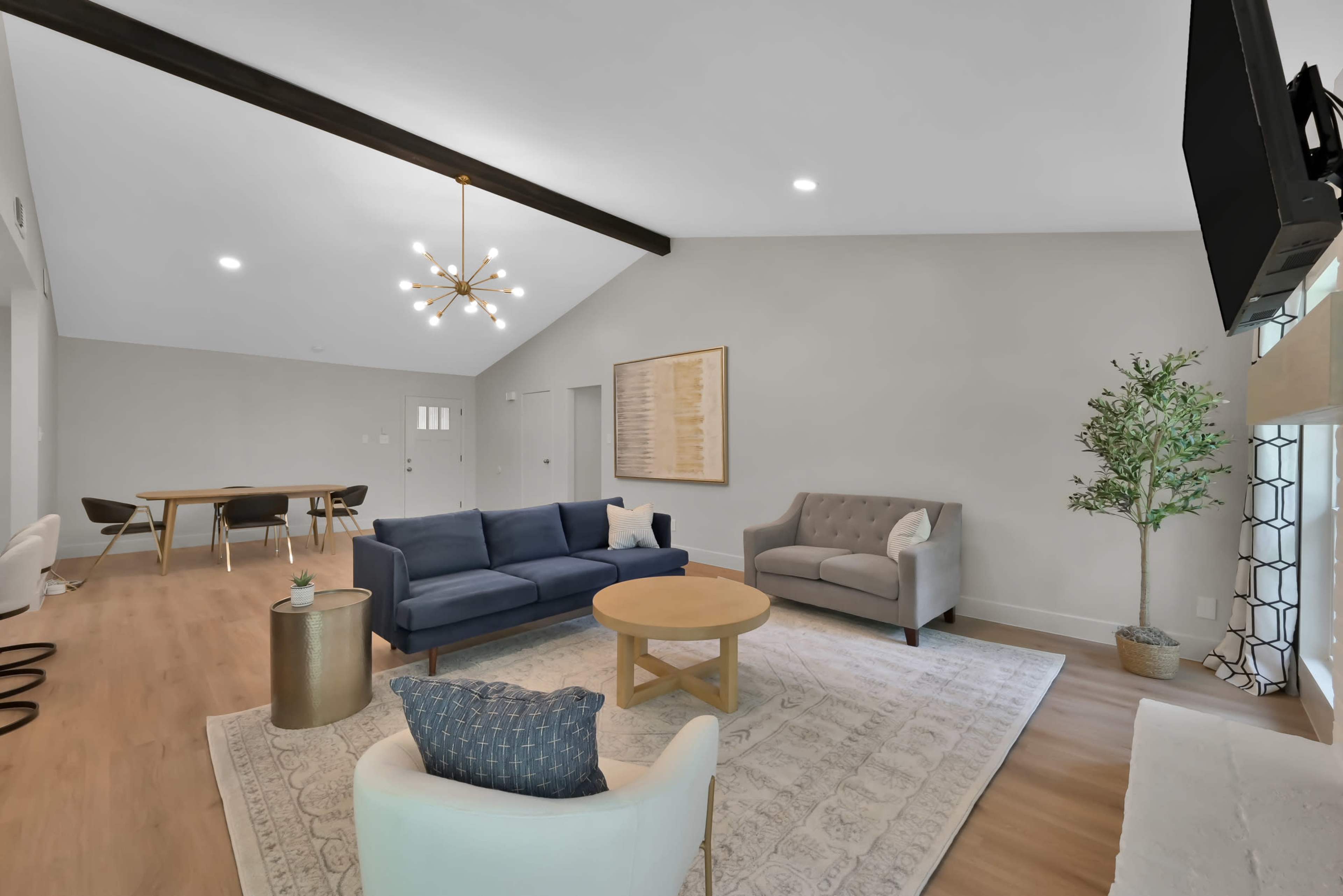 A modern living room featuring a blue sofa, a light gray couch, a round wooden coffee table, and a decorative plant, with a dining area visible in the background.
