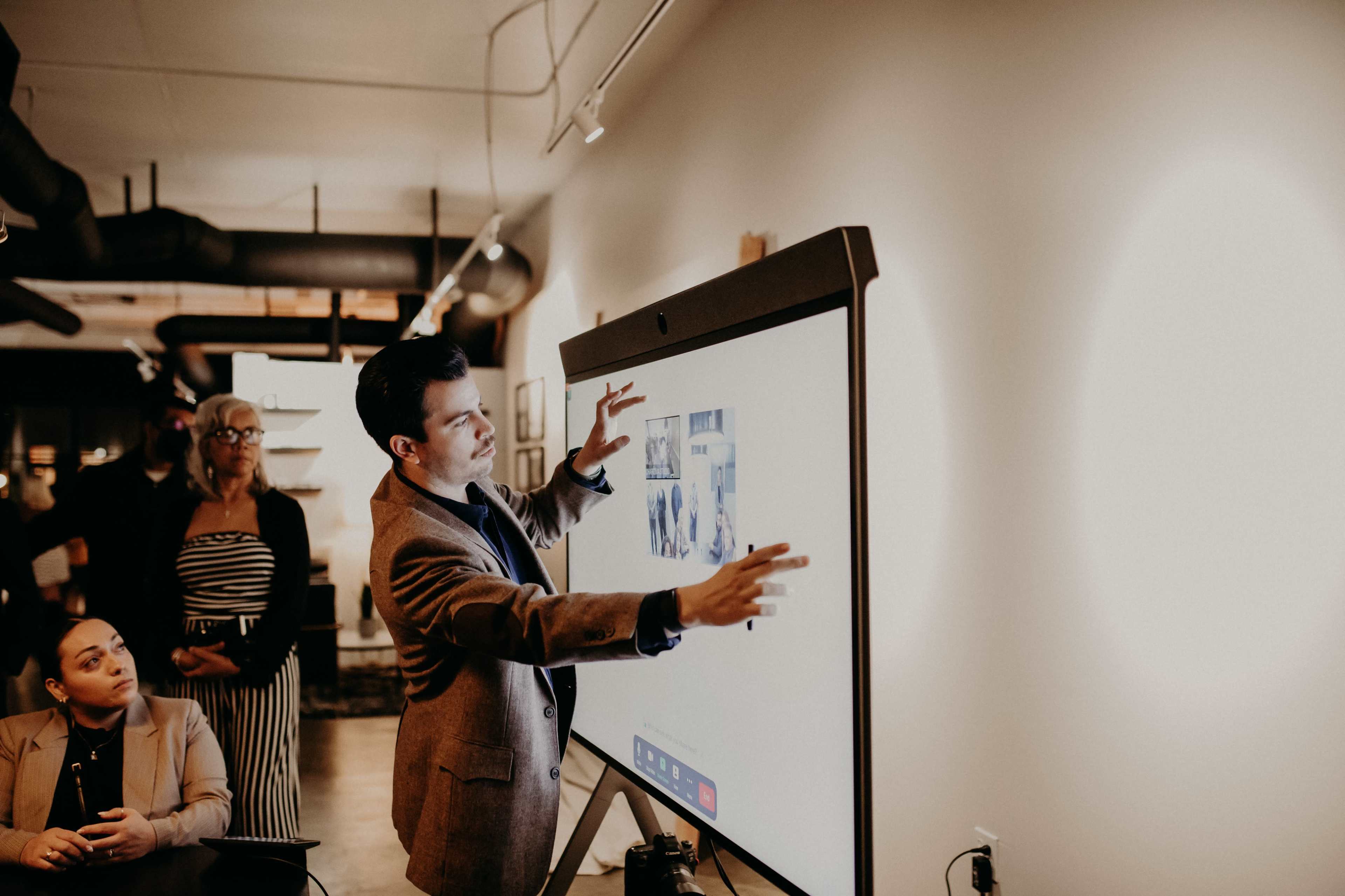 A man is presenting on a touchscreen display while an audience watches attentively in a modern office setting.