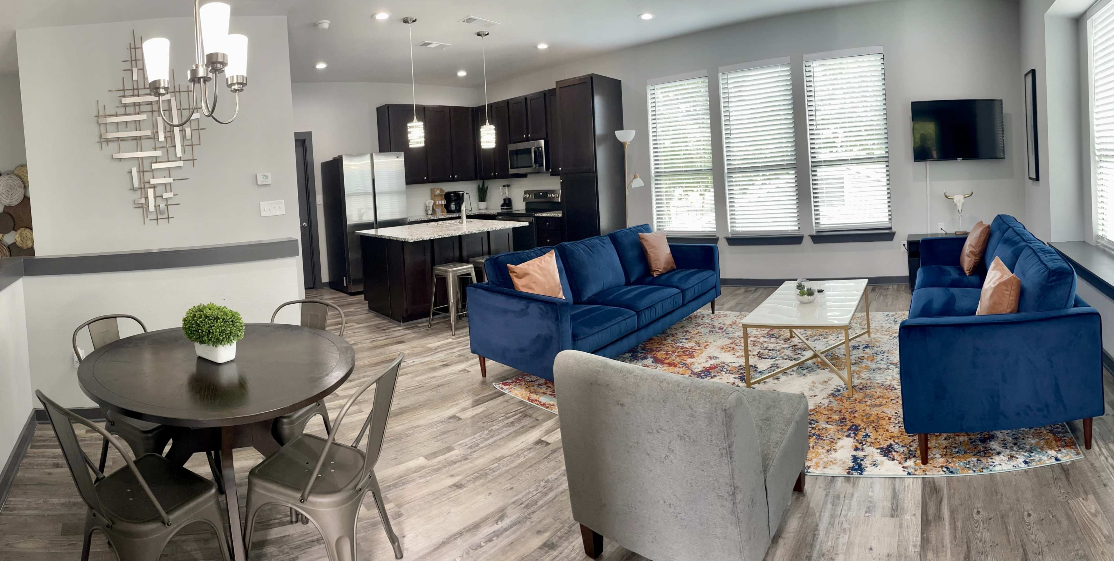 A modern living space featuring a blue sectional sofa, a round dining table, and a kitchen area with dark cabinetry and stainless steel appliances.